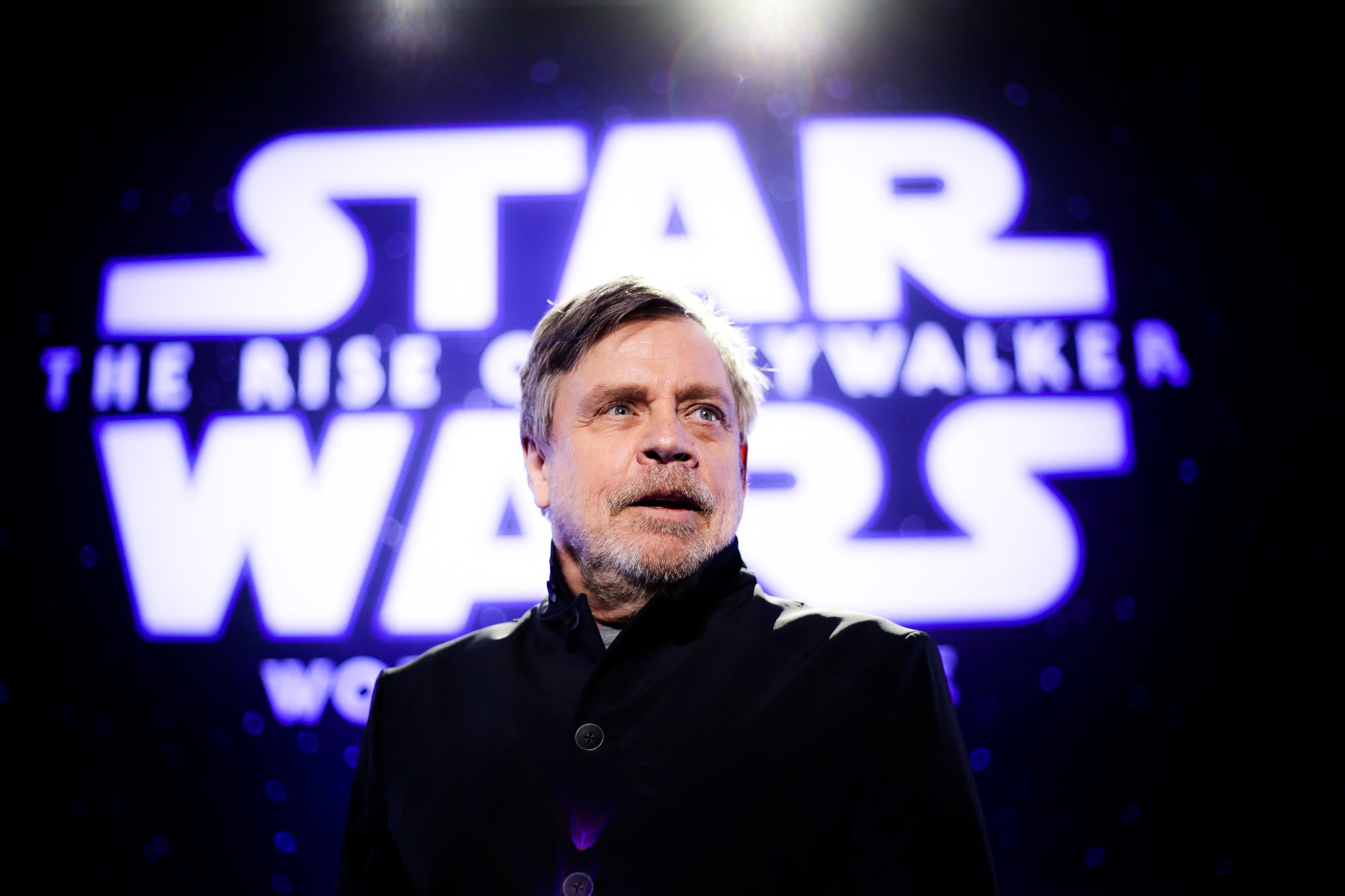'Star Wars' actor Mark Hamill standing in front of lit up logo wearing a black coat