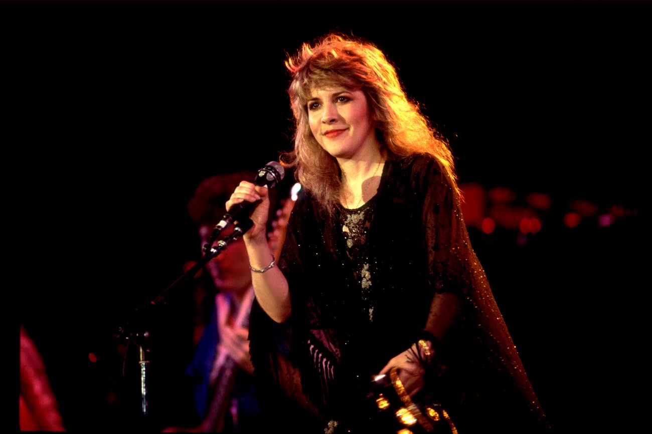 Stevie Nicks wears a black shawl and stands onstage with a microphone. 