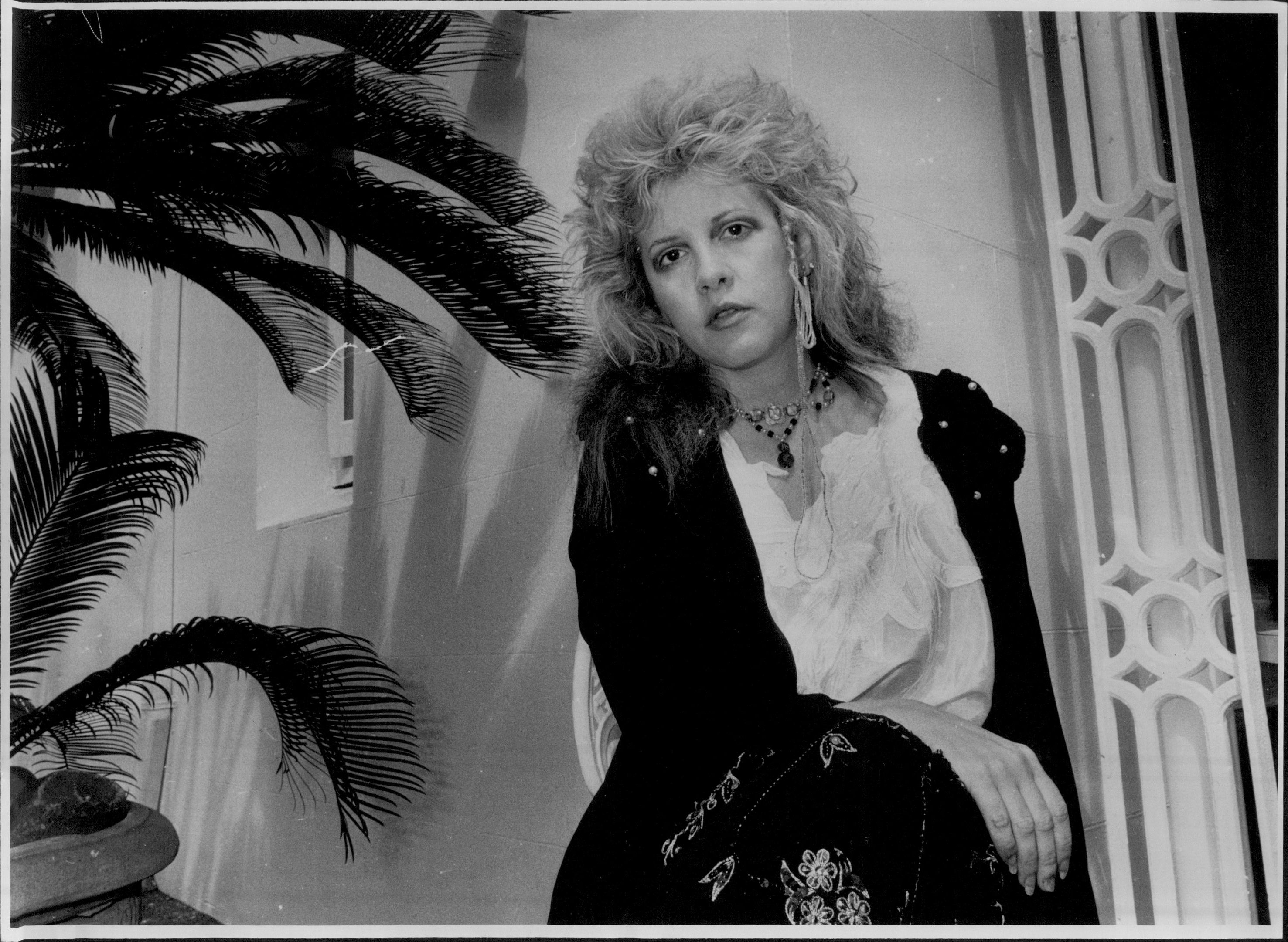 A black and white picture of Stevie Nicks sitting near a potted palm plant in 1986, the same year she went to rehab.