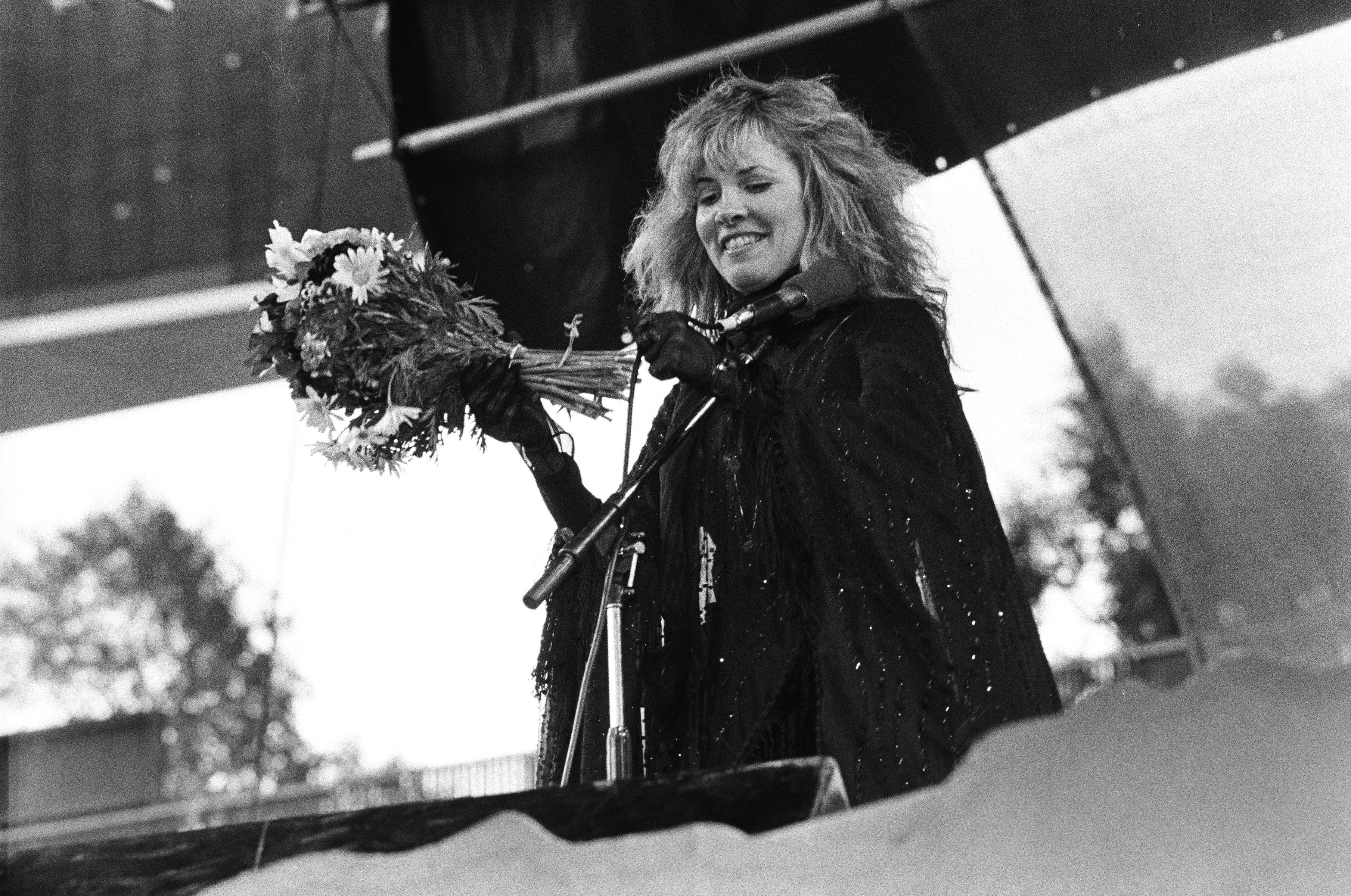 A black and white photo of Stevie Nicks wearing a black dress and holding a bouquet of flowers from a fan.