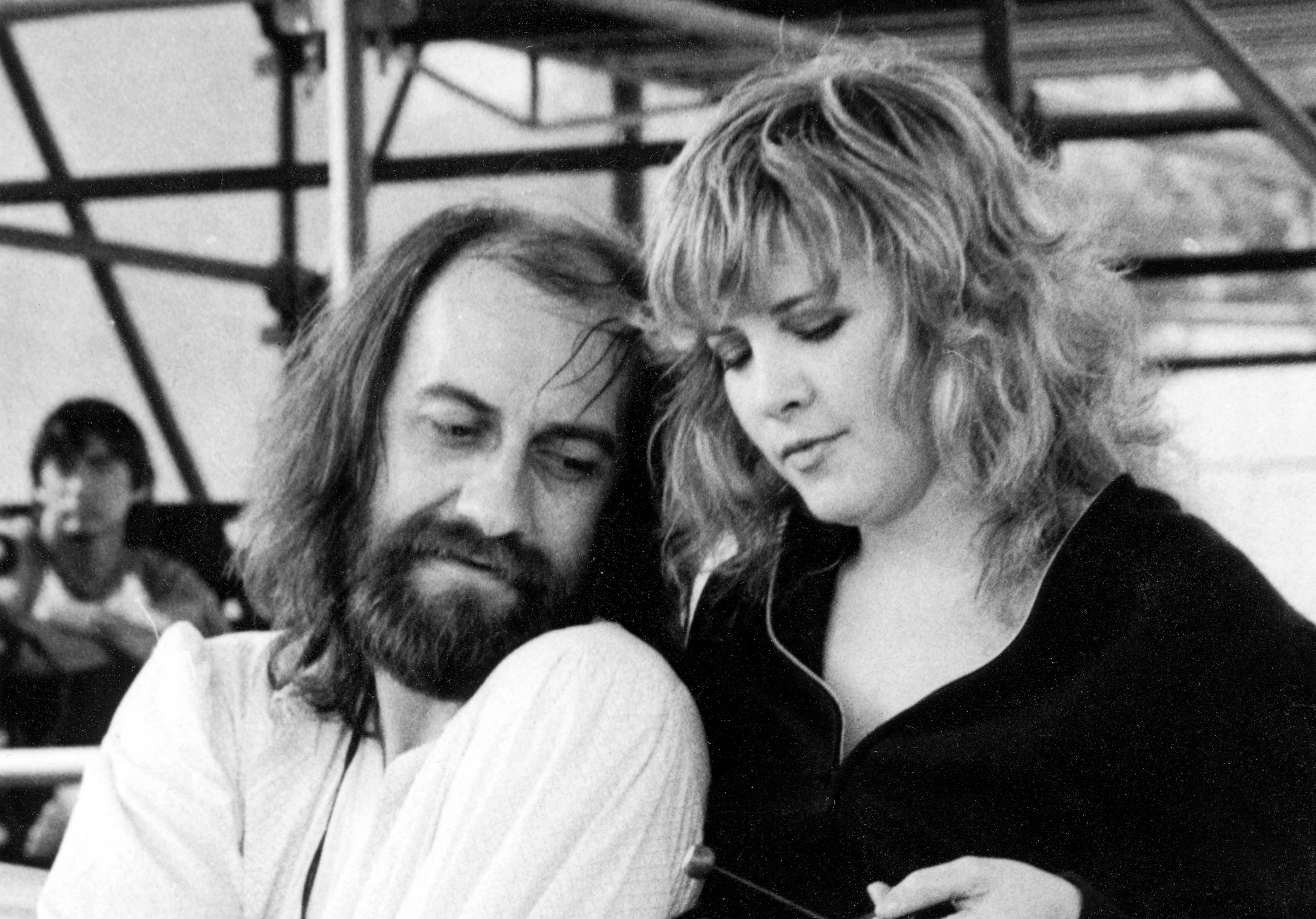 A black and white picture of Mick Fleetwood and Stevie Nicks. Mick Fleetwood sits while Stevie Nicks stands behind him.