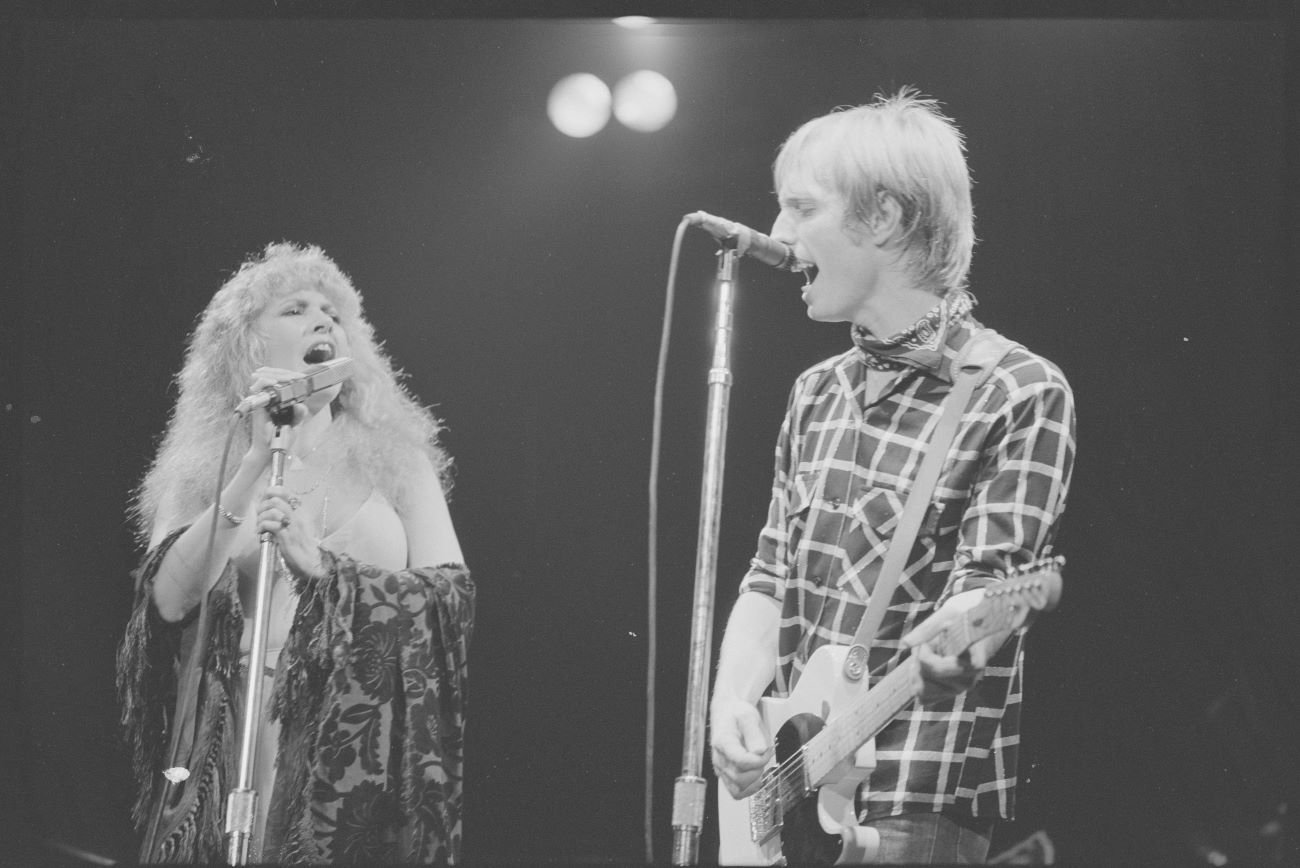 A black and white photo of Stevie Nicks and Tom Petty on stage together singing into microphones.