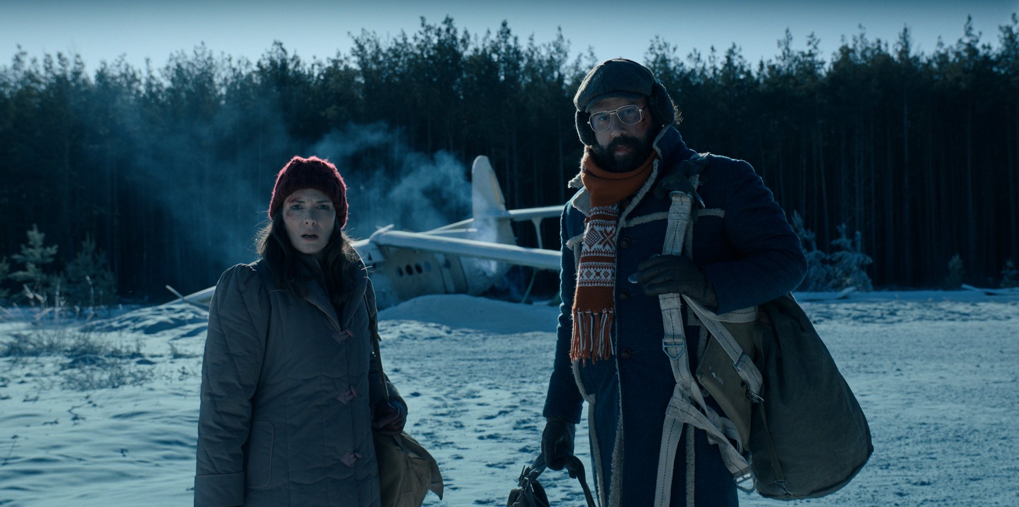 'Stranger Things 4' stars Brett Gelman and Winona Ryder in front of a crashed airplane on a snowy landscape in a production still from the series.