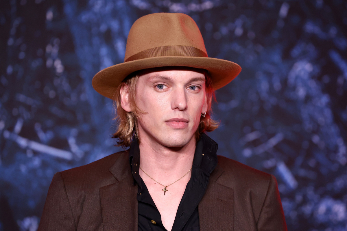 Jamie Campbell Bower attends the Stranger Things Season 4 premiere wearing a tan hat, brown jacket and black shirt. 