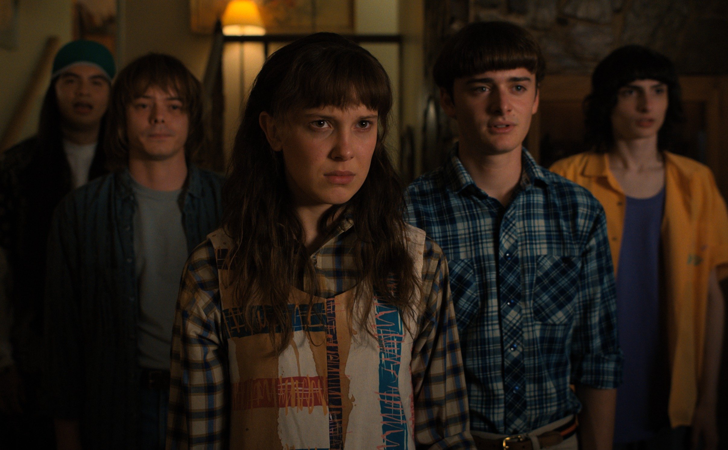 Eduardo Franco, Charlie Heaton, Millie Bobby Brown, Noah Schnapp, Finn Wolfhard in 'Stranger Things 4.' whiich set its part 2 release date for July. The group is standing in a dimly lit room and looking skeptically at someone or something off-screen.