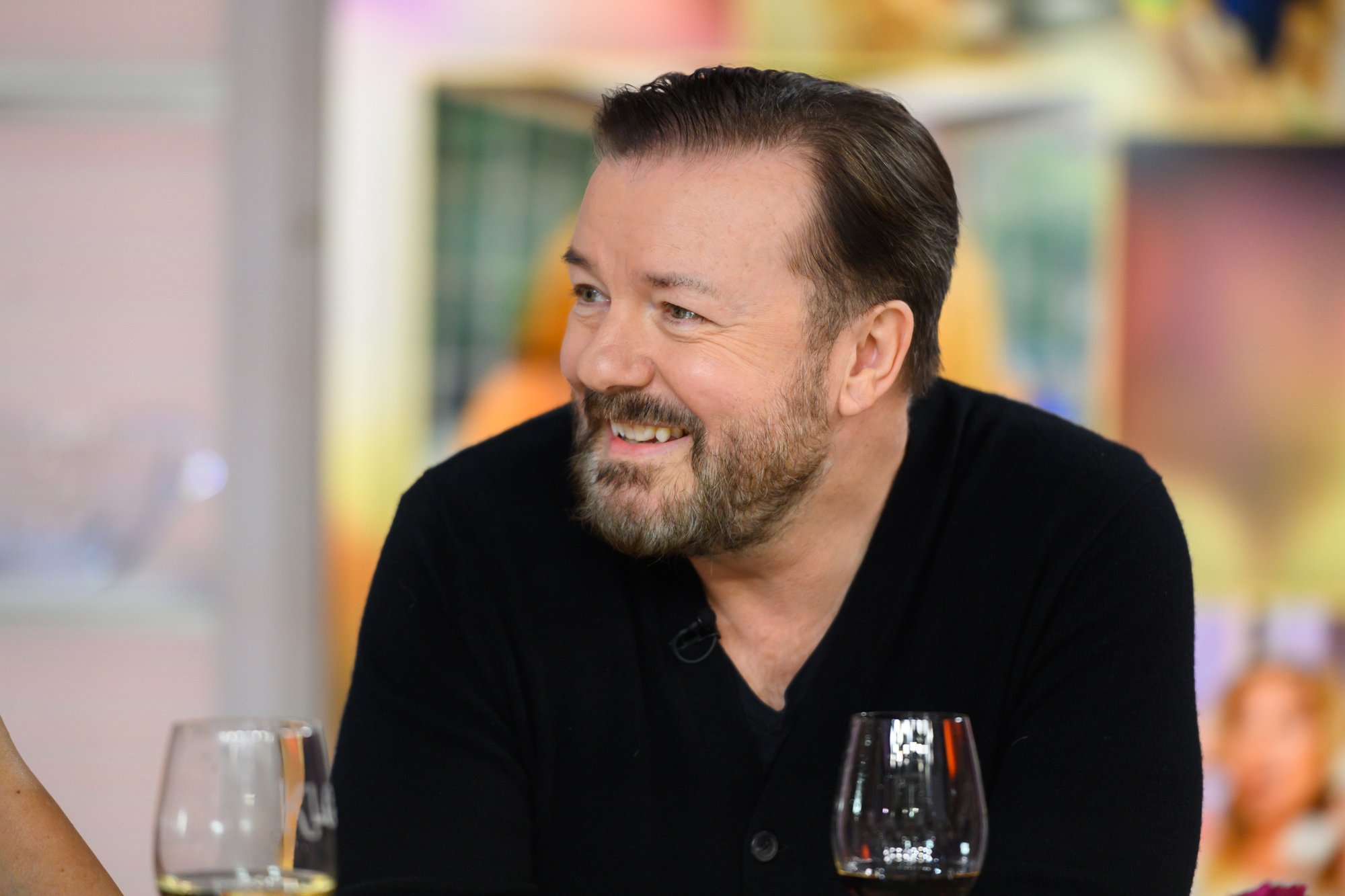 'SuperNature' comedian Ricky Gervais smiling and wearing a black shirt with a glass of wine in front of him