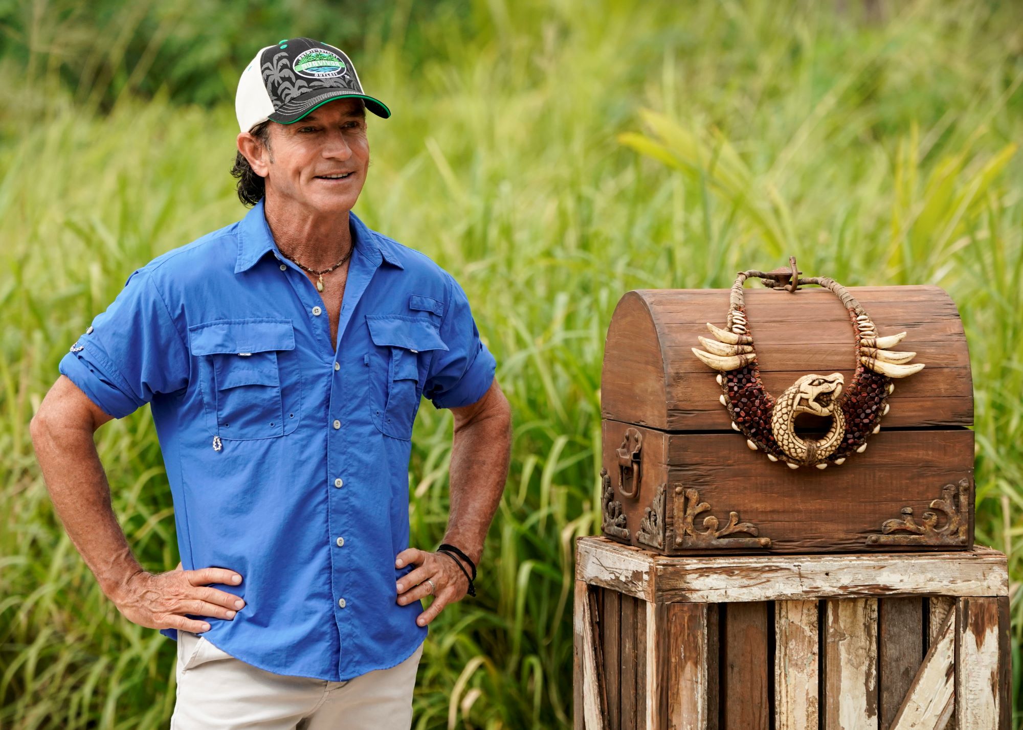 Jeff Probst, who hosts 'Survivor' Season 42 on CBS, wears a blue short-sleeve button-up shirt, light pants, and a black, white, and green 'Survivor' baseball cap.
