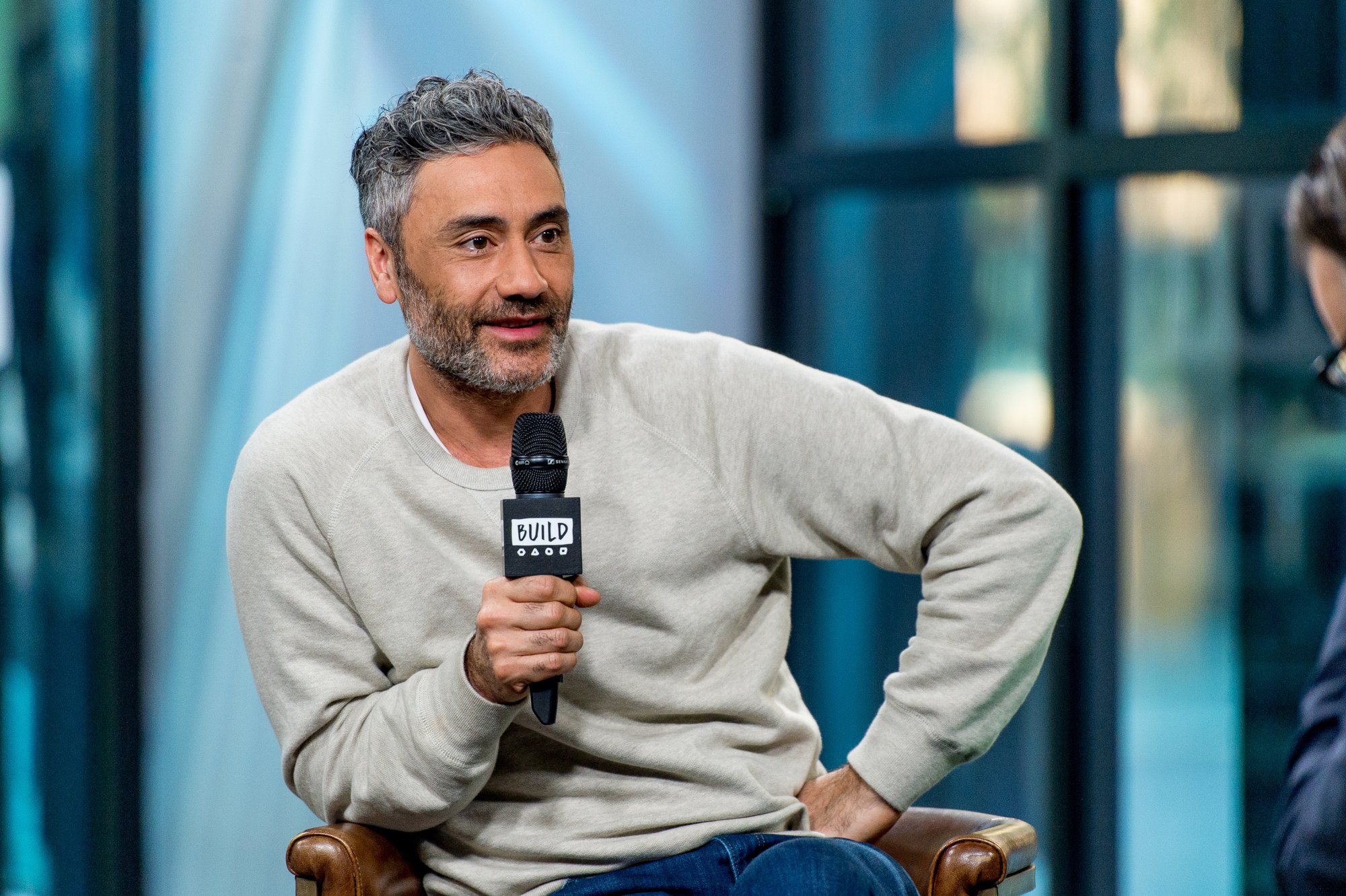 Taika Waititi, the director who decided to bring Jane Foster back for 'Thor: Love and Thunder.' He's sitting down and holding a microphone, and he looks like he's about to say something.