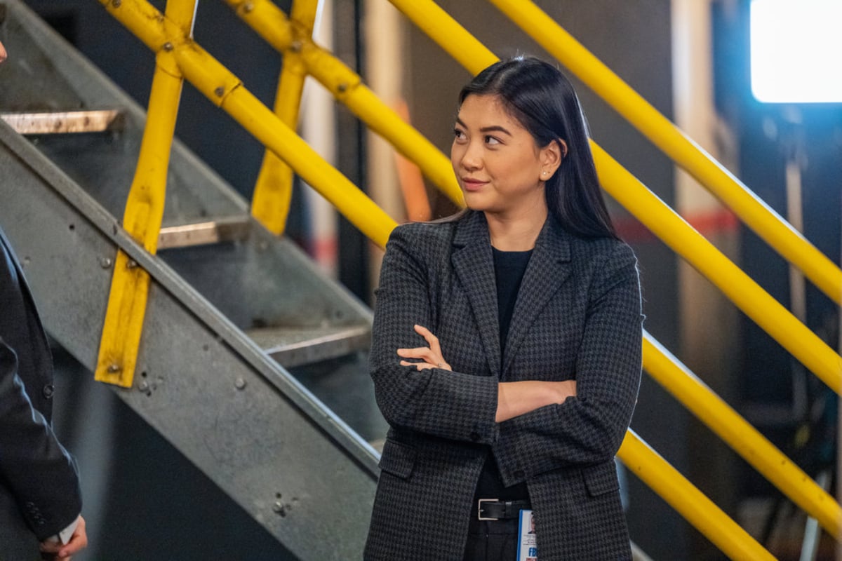 Laura Sohn as Alina Park in The Blacklist Season 9. Park stands in the Post Office with her arms crossed, smiling. 