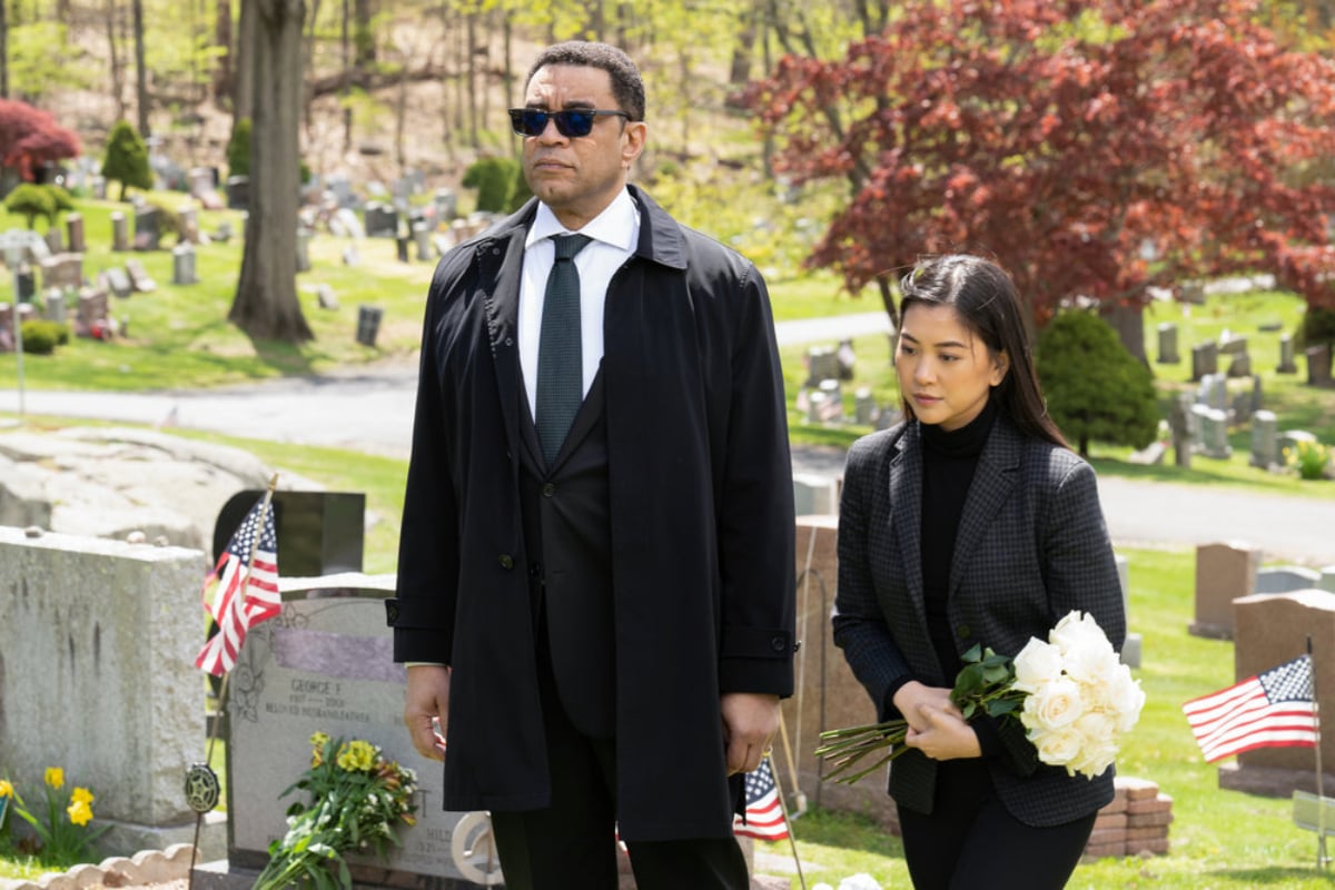 Cooper and Park stand in a cemetery in 'The Blacklist' Season 9 Episode 22. Park holds flowers and Cooper wears sunglasses.