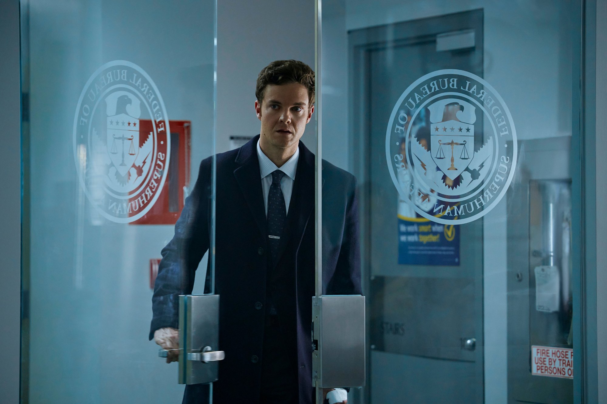 Jack Quaid as Hughie Campbell in 'The Boys' Season 3. He's wearing a suit and walking through a set of glass doors.