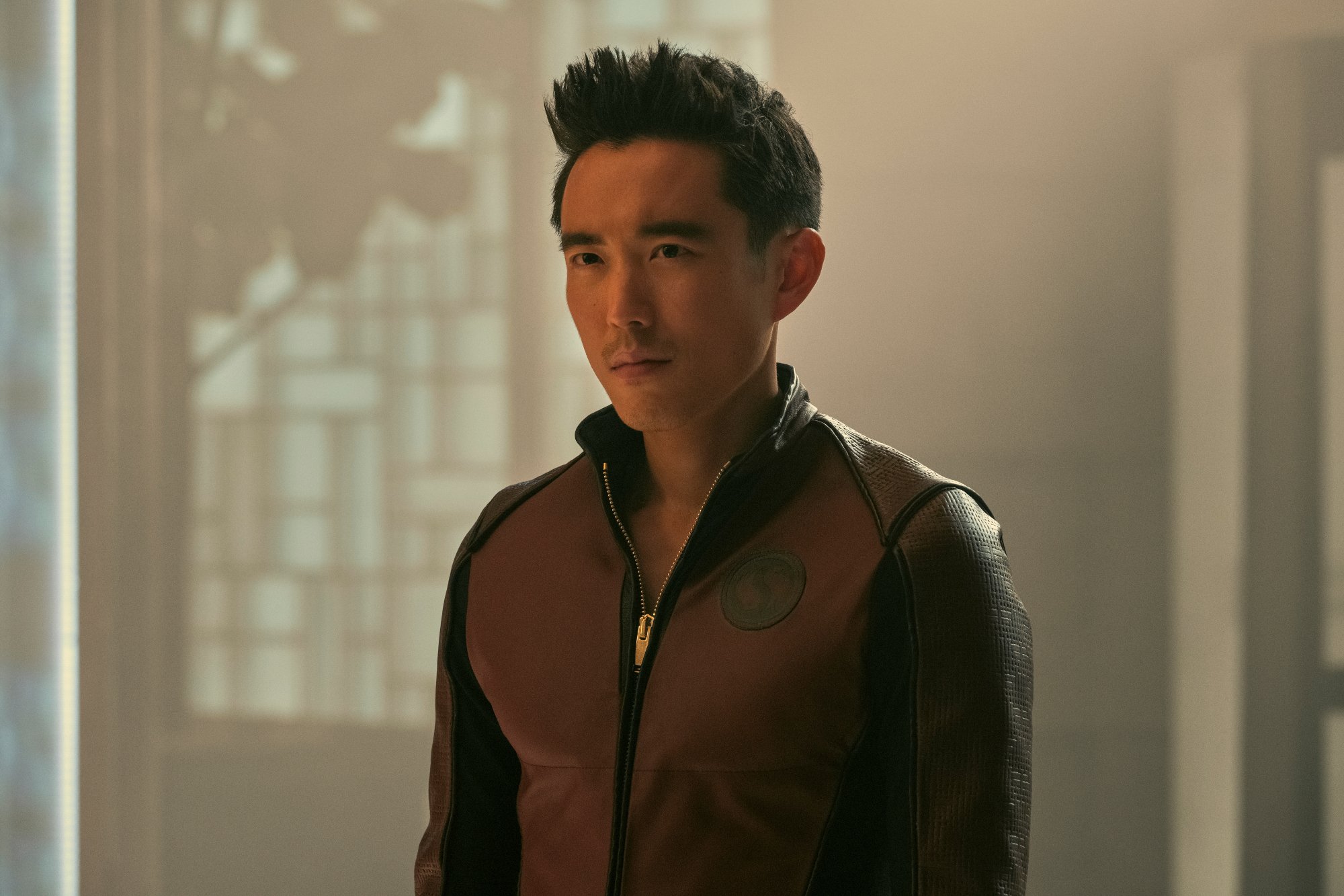 Justin H. Min as Ben Hargreeves in 'The Umbrella Academy' Season 3 on Netflix. He's wearing a red and black jacket, and his hair is spiked up.