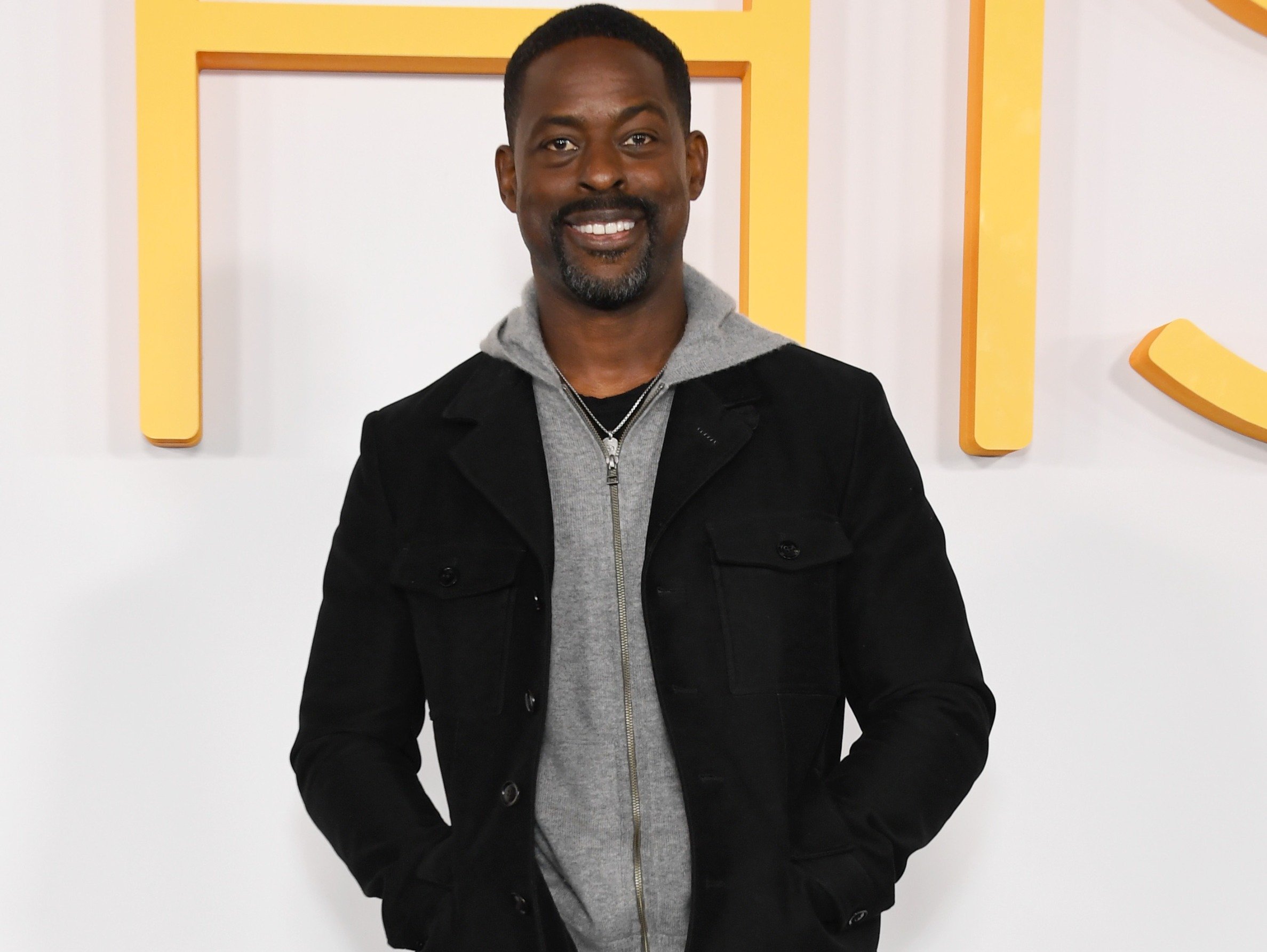 'This Is Us' star Sterling K. Brown, who appears as Randall Pearson for the last time in the series finale. He's standing in front of a white wall with the yellow 'This Is Us' logo on it, and he's wearing a gray sweatshirt under a black jacket.