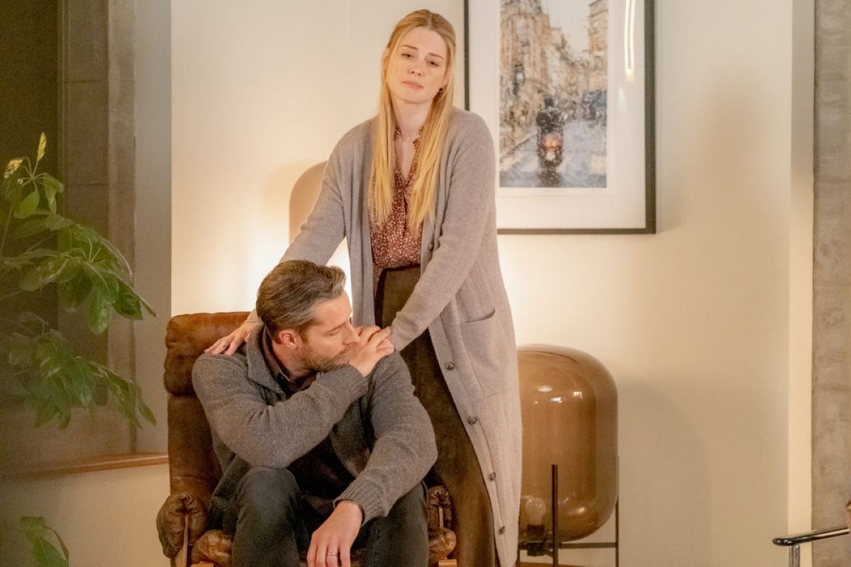Justin Hartley and Alexandra Breckenridge, in character as Kevin and Sophie in 'This Is Us' Season 6 Episode 17, share a scene where Kevin sits on a chair and Sophie stands behind him with her hands on his shoulders. Kevin wears a gray sweater and jeans. Sophie wears a long gray cardigan over a red and white blouse and brown skirt.