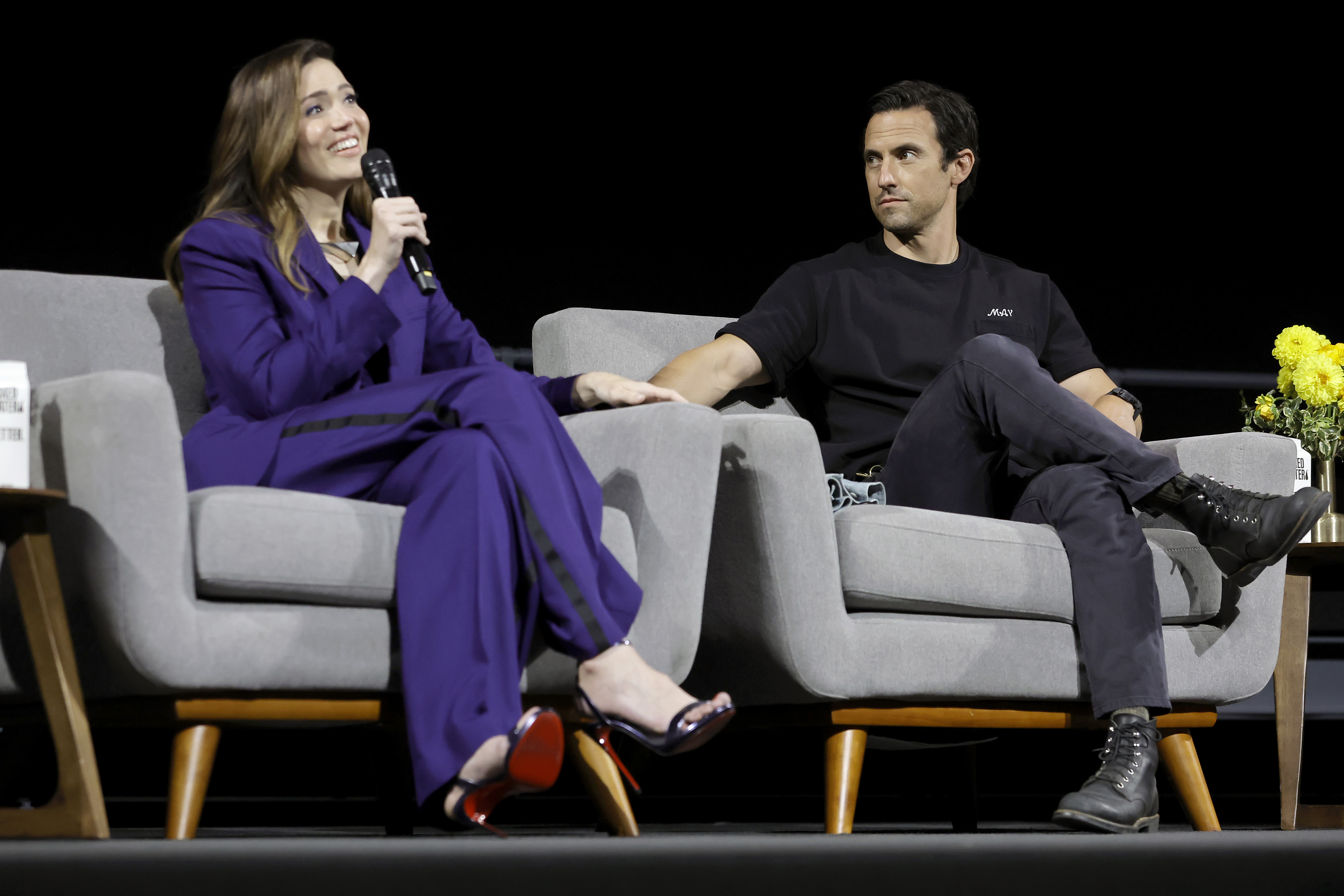 Mandy Moore and Milo Ventimiglia, who played Rebecca and Jack in 'This Is Us' Season 6, sit next to each other onstage. Moore wears a dark blue suit. Ventimiglia wears a black shirt and gray pants.