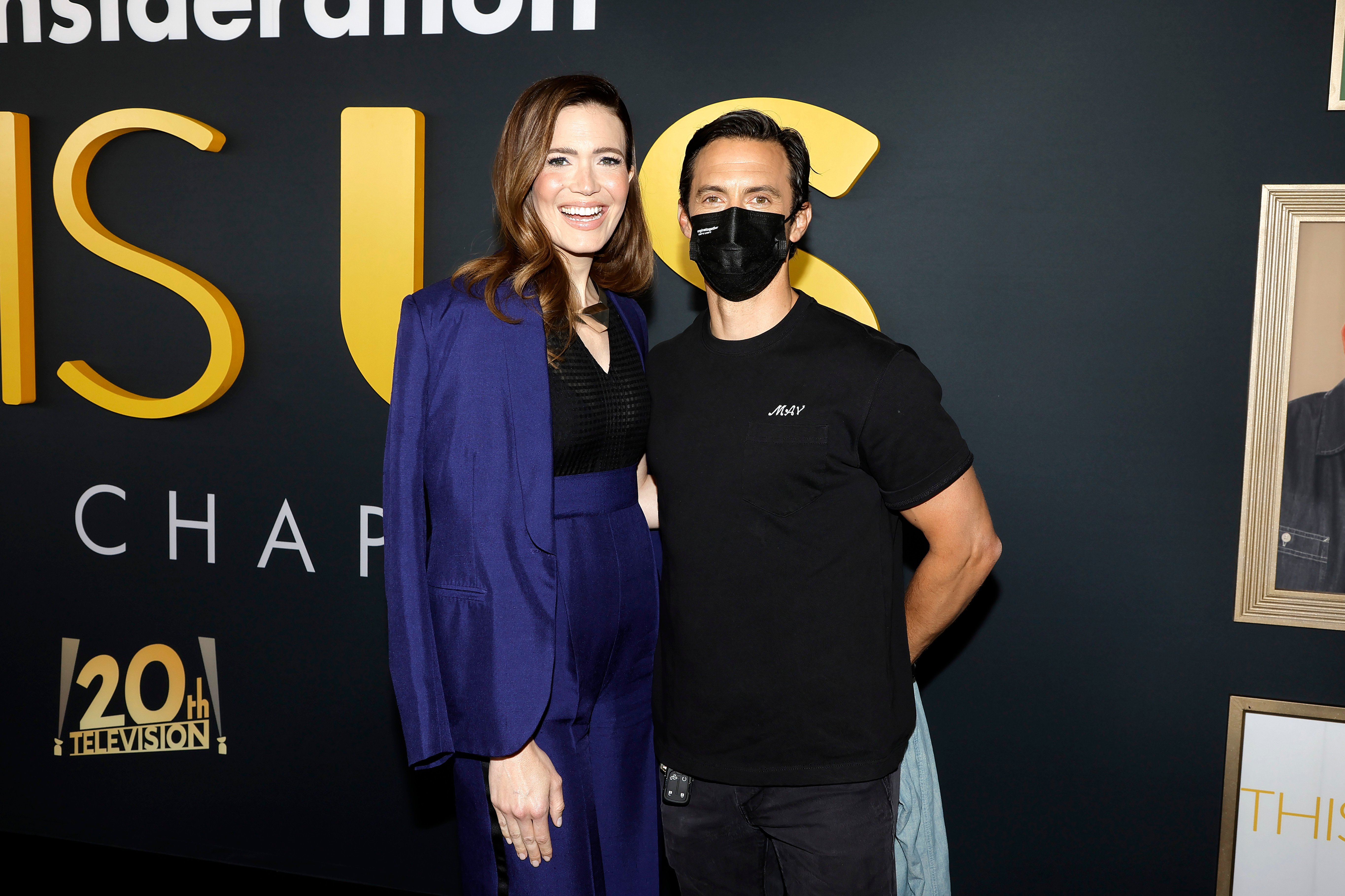 Mandy Moore and Milo Ventimiglia, who star as Rebecca and Jack in 'This Is Us' Season 6, pose for pictures together. Moore wears a dark blue suit over a black shirt and dark blue pants. Ventimiglia wears a black shirt and black pants.