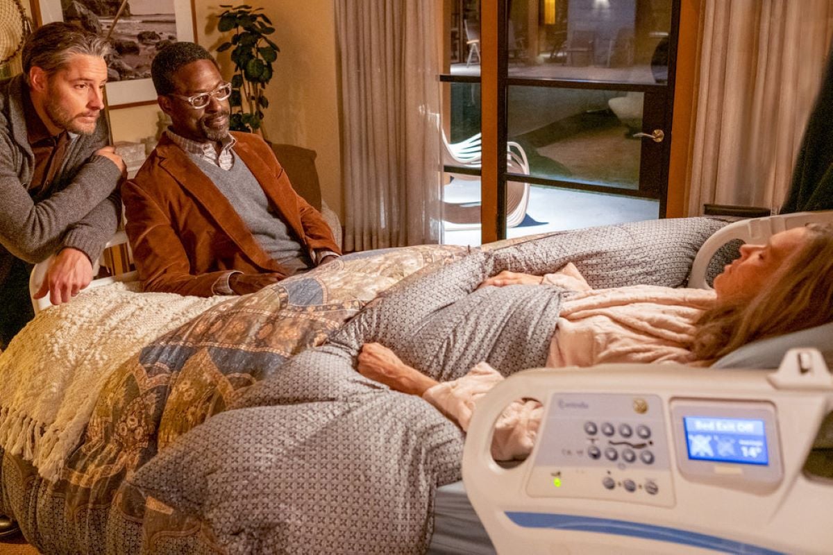 Justin Hartley as Kevin, Sterling K. Brown as Randall, and Mandy Moore as Rebecca share a scene in 'This Is Us' Season 6 Episode 17. Rebecca, in a pink robe, lays in bed while Kevin, wearing a gray cardigan, and Randall, wearing a brown jacket over a gray sweater, look at her.