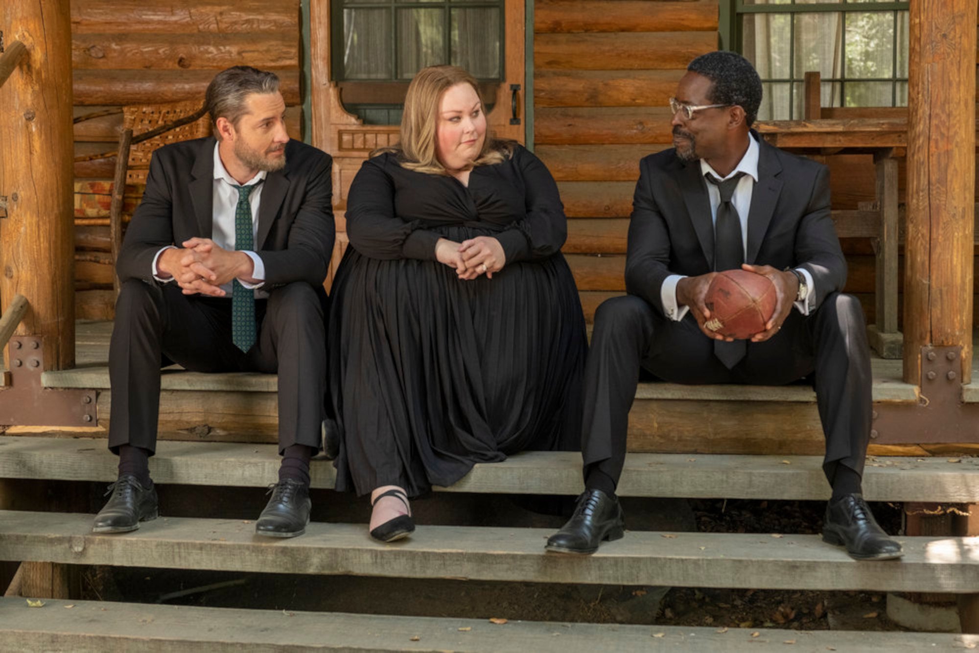 Justin Hartley as Kevin, Chrissy Metz as Kate, Sterling K. Brown as Randall in 'This Is Us' Season 6 Episode 18. One 'This Is Us' theory posits the Big 3 will be affected by Dr. Marcus Brooks' research. In this photo, they're sitting on the porch at Rebecca's funeral. They're wearing black and talking to one another.