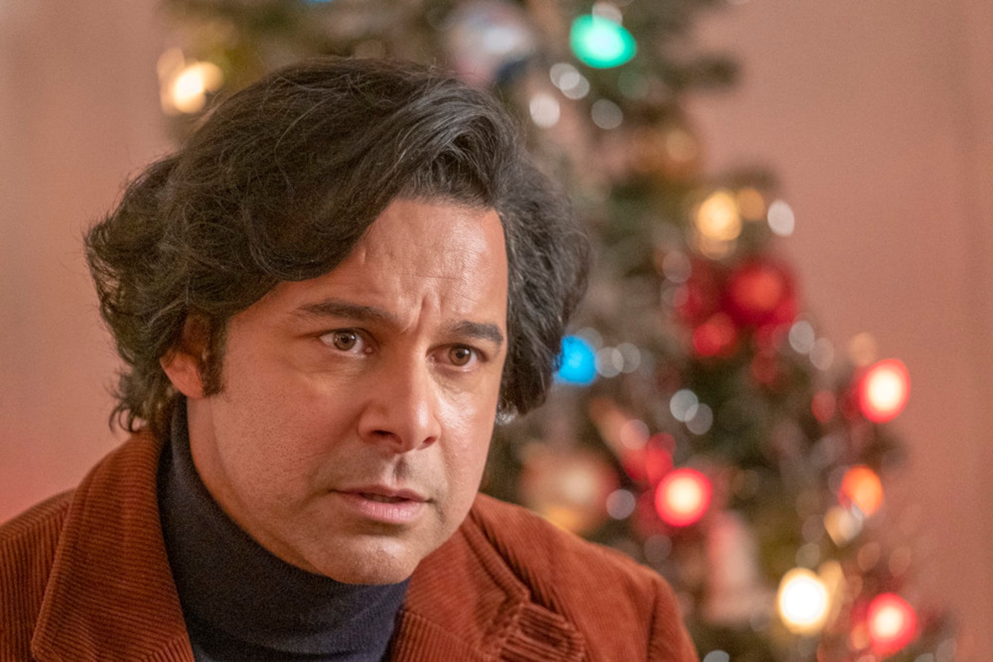 Jon Huertas as Miguel in 'This Is Us' Season 6 Episode 15. He's wearing black turtle neck and orange vest and sitting in front of a Christmas tree.