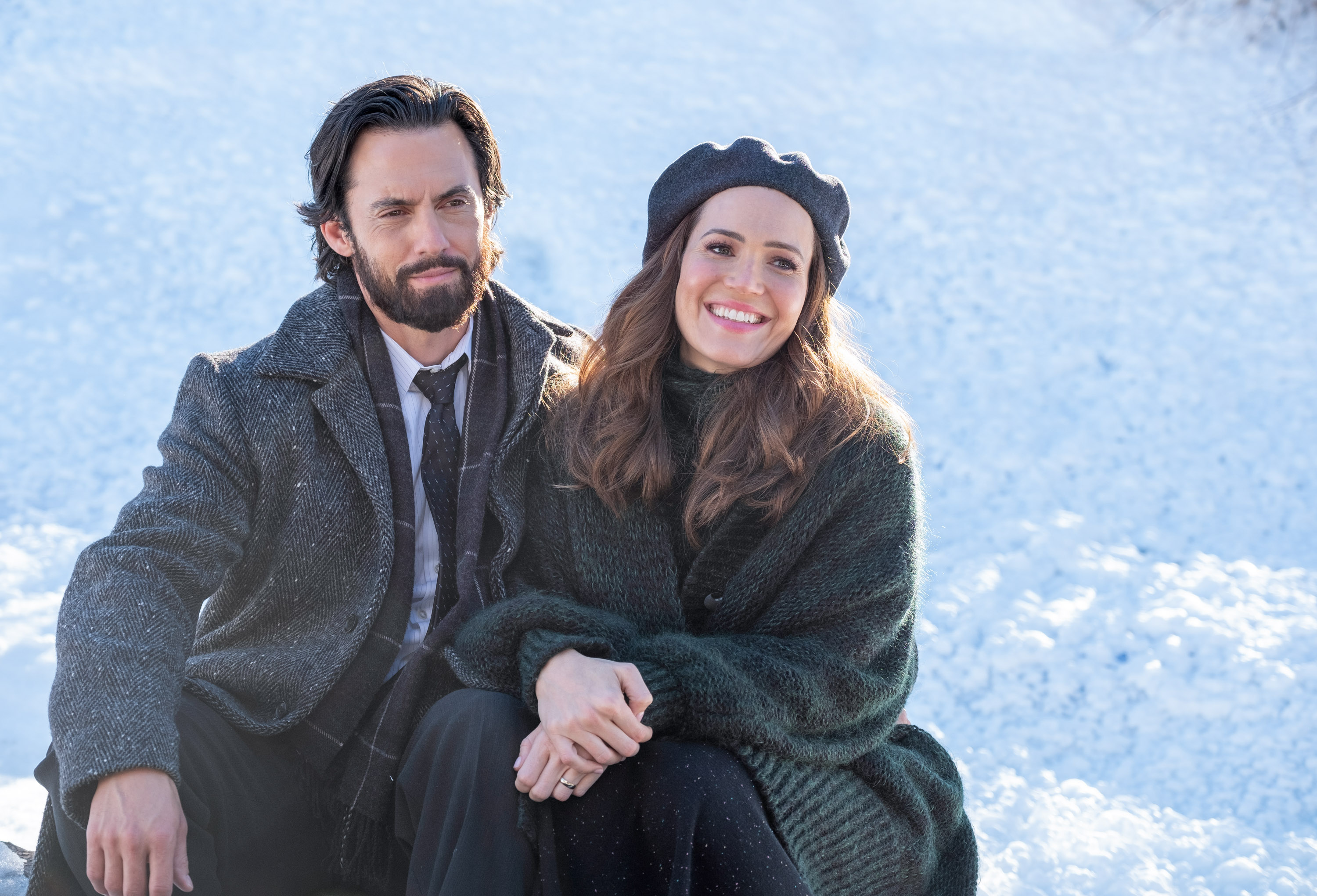 Milo Ventimiglia and Mandy Moore, in character as Jack and Rebecca in 'This Is Us' Season 6 Episode 4, share a scene. Jack wears a gray coat over a white button-up shirt, black tie, and black pants. Rebecca wears a oversized gray sweater, a long black skirt, and a black hat.