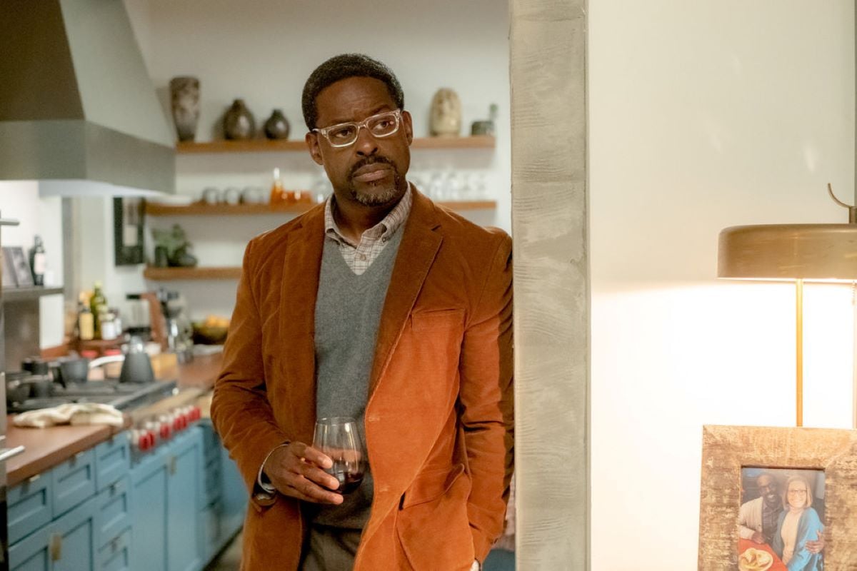 Sterling K. Brown, in character as Randall in 'This Is Us' Season 6 Episode 17, wears clear glasses and a light brown suit over a light gray sweater vest over a light brown plaid shirt while holding a glass of wine.