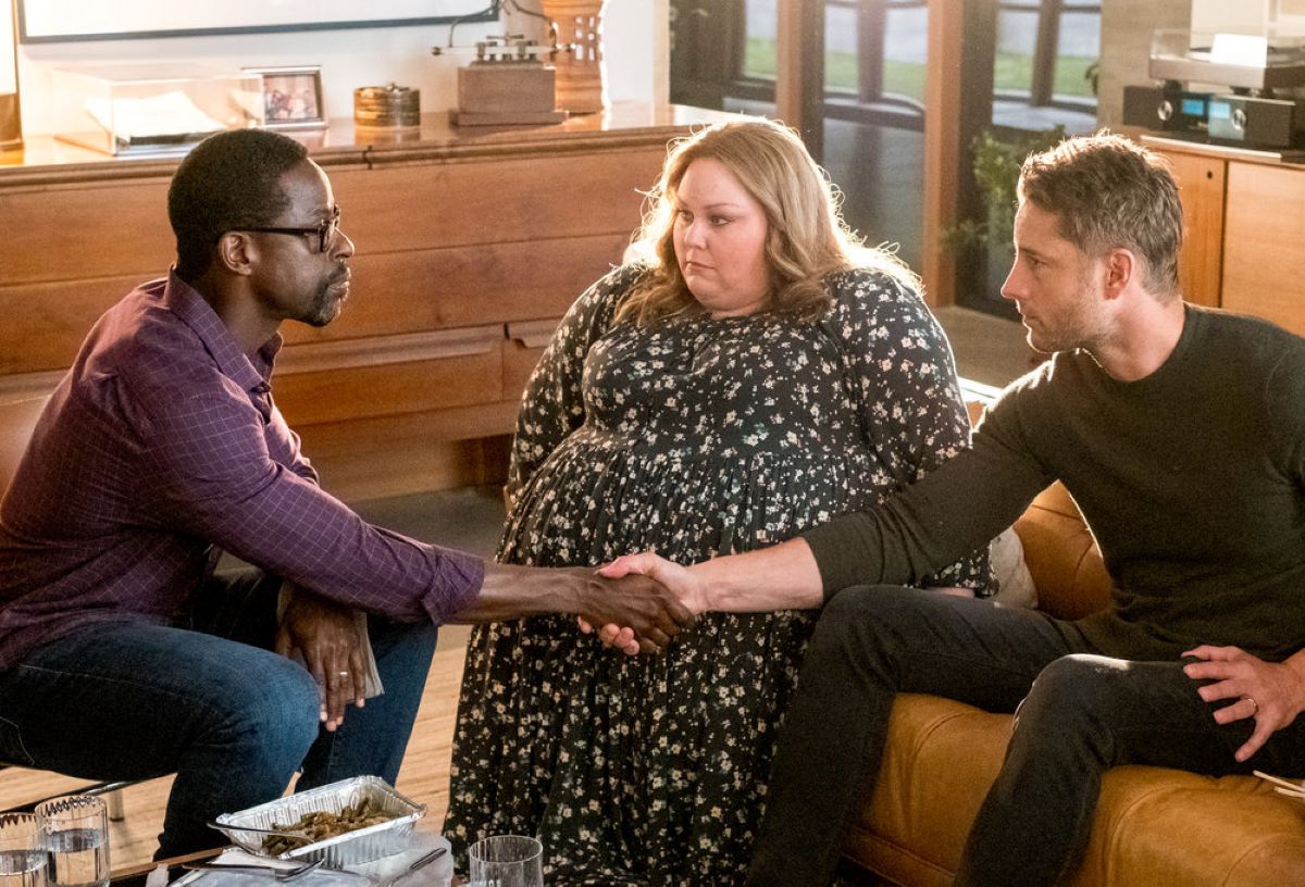 'This Is Us' Season 6 Episode 16 stars Sterling K. Brown, Chrissy Metz, and Justin Hartley, in character as Randall, Kate, and Kevin, share a scene. Randall and Kevin shake hands, and Kate sits between them. Randall wears a maroon long-sleeved button-up shirt and jeans. Kate wears a black dress with white flowers on it. Kevin wears a dark green sweater and black pants.