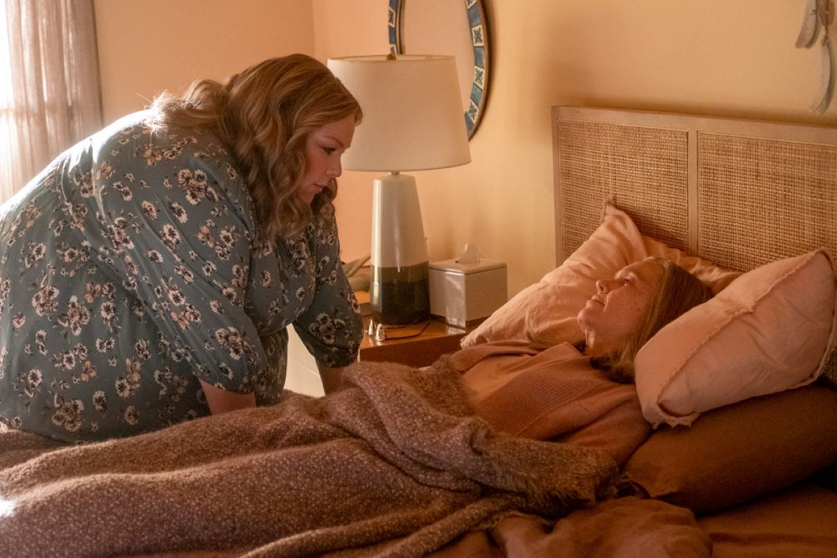 'This Is Us' Season 6 Episode 17 stars Chrissy Metz and Mandy Moore, in character as Kate and Rebecca, share a scene where Kate tucks Rebecca into bed. Kate wears a blue dress with white flowers on it. Rebecca wears a pink cardigan over a white shirt.