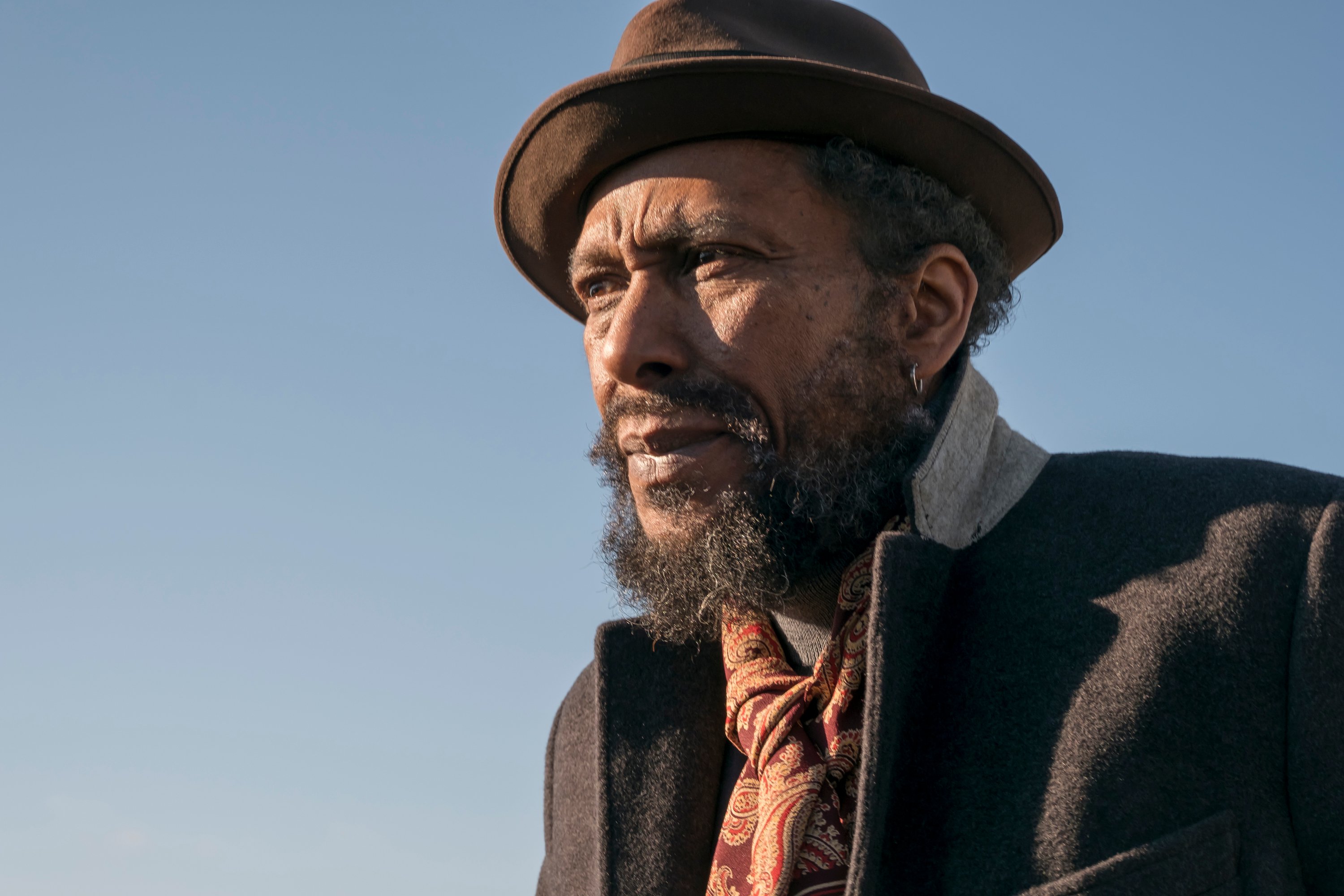 Ron Cephas Jones, in character as William in 'This Is Us' Season 1, wears a black coat, a red and gold paisley scarf, and a brown hat.