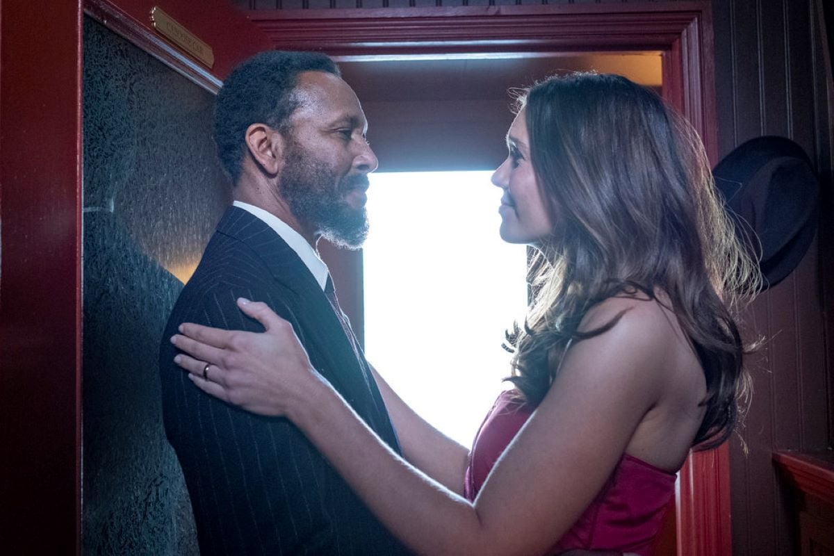 Ron Cephas Jones and Mandy Moore, in character as William and Rebecca in 'This Is Us' Season 6 Episode 17, share a scene where they look at one another and Rebecca has her hands on William's shoulders. William wears a black suit with thin white stripes. Moore wears a red dress.