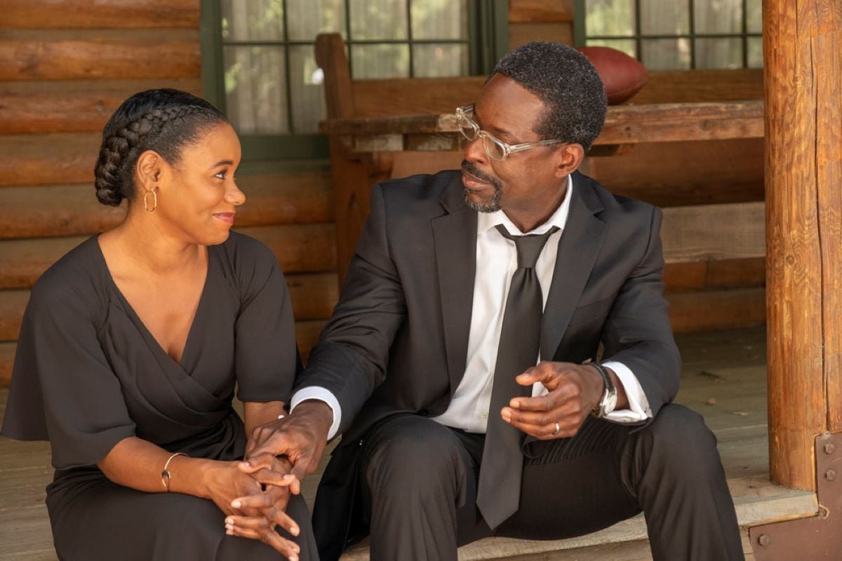 La Trice Harper and Sterling K. Brown, in character as Deja and Randall in 'This Is Us' Season 6 Episode 18, share a scene on porch steps while Randall holds Deja's hands. Deja wears a black dress. Randall wears a black suit over a white button-up shirt and black tie.