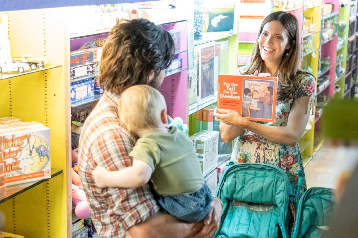 Milo Ventimiglia and Mandy Moore, in character as Jack and Rebecca, share a scene in the final episode of their show, 'This Is Us' Season 6 Episode 18. Jack, while holding a baby, wears an orange, white, and brown plaid shirt. Rebecca, while holding a game of pin the tail on the donkey, wears a white, red, green, and yellow floral dress.