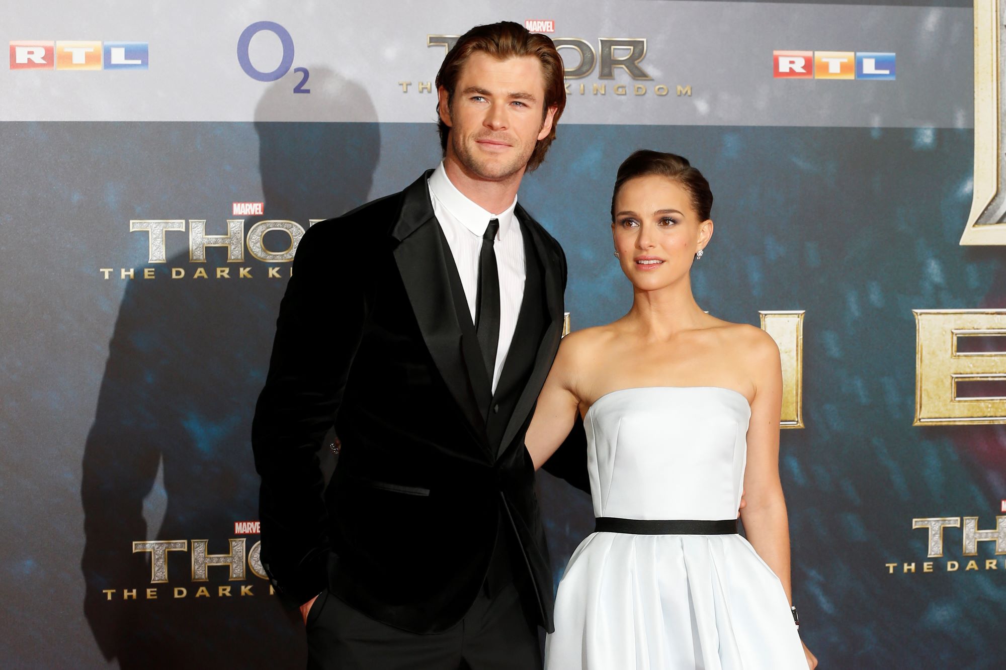 Chris Hemsworth and Natalie Portman, who star as Thor and Jane in the 'Thor: Love and Thunder' trailer, pose for pictures together. Hemsworth wears a black velvet suit over a white button-up shirt and black tie. Portman wears a white strapless dress with a thin black ribbon around the waist.
