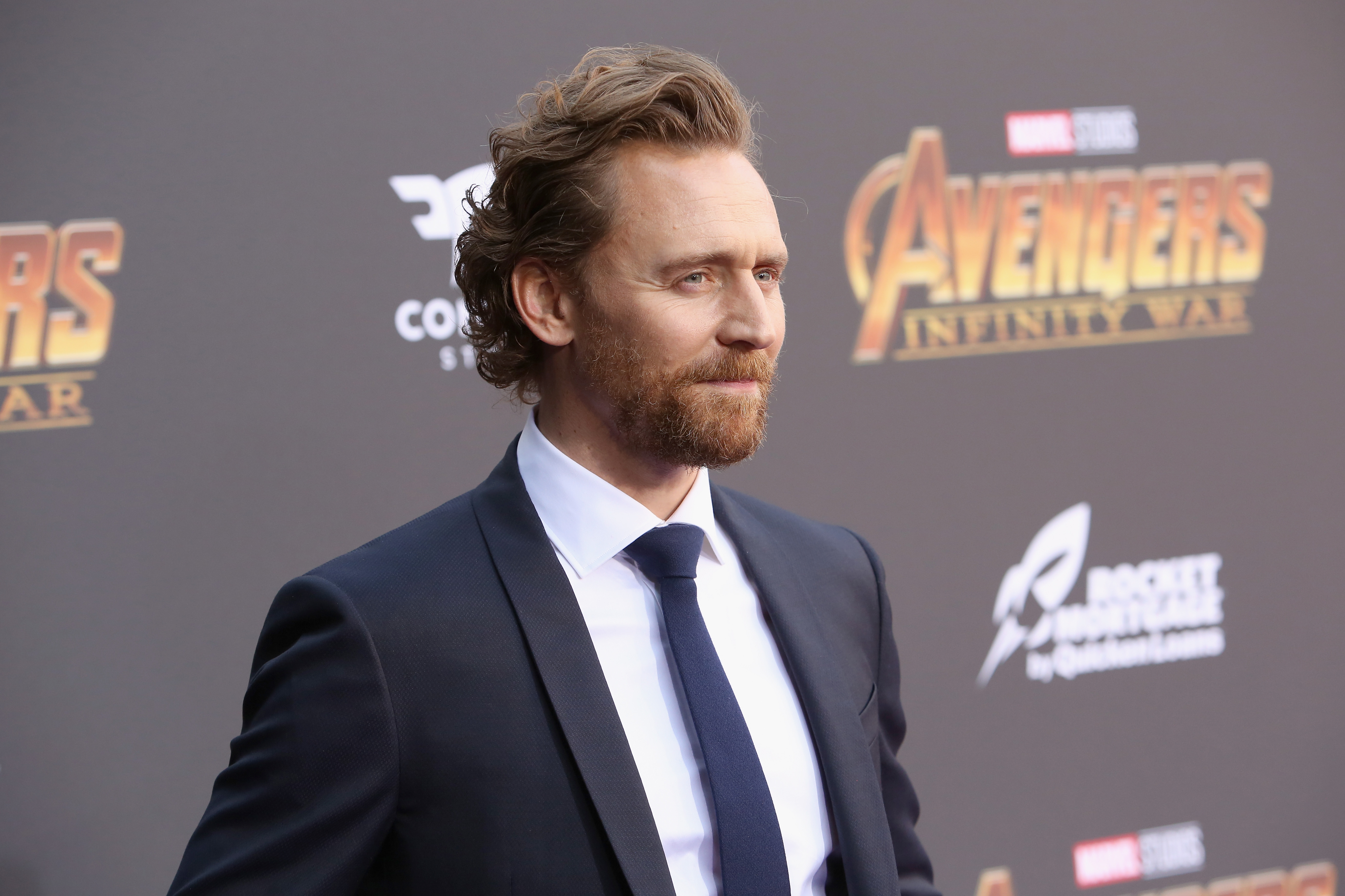 Tom Hiddleston, who might appear in 'Thor: Love and Thunder' as Loki, wears a black suit over a white button-up shirt and black tie.