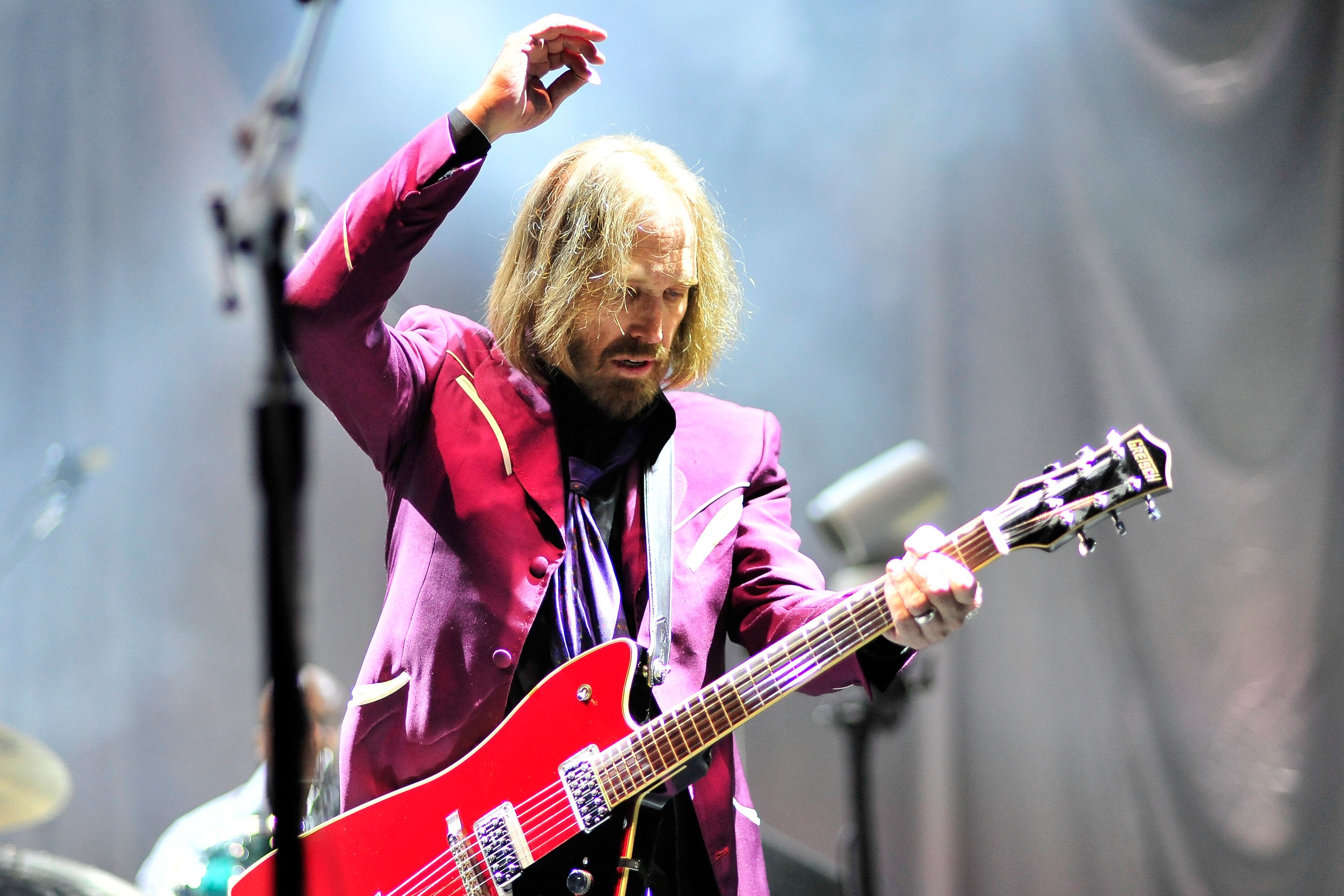 Tom Petty wears a purple jacket and holds his hand in the air while playing guitar.