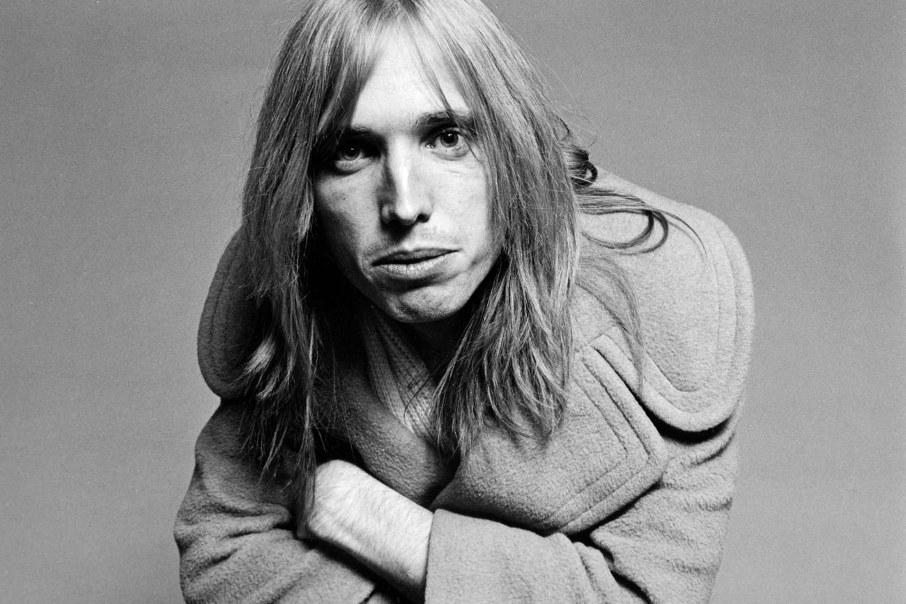 A black and white photo of Tom Petty leaning down to look at the camera with his arms folded.