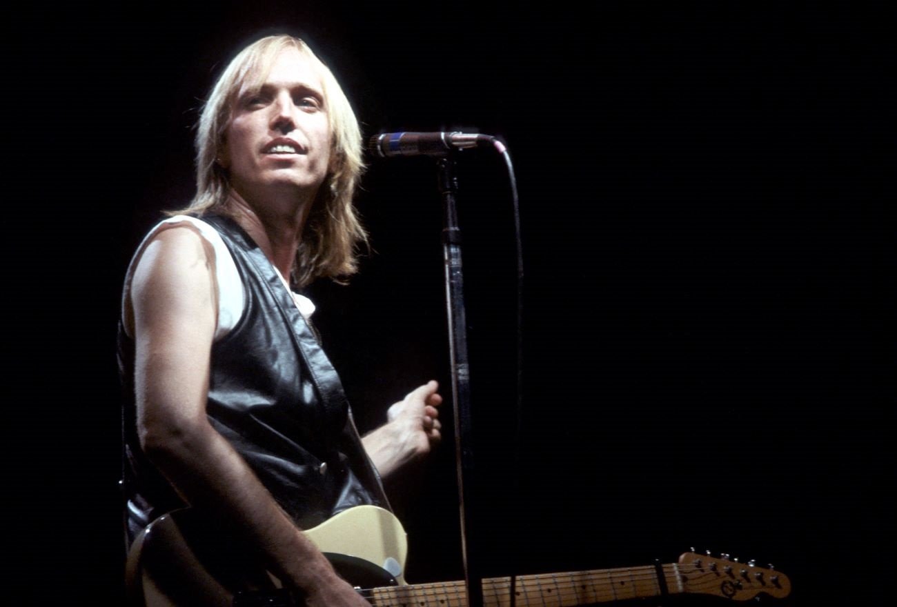 Tom Petty wears a tank top, holds a guitar, and stands in front of a microphone.