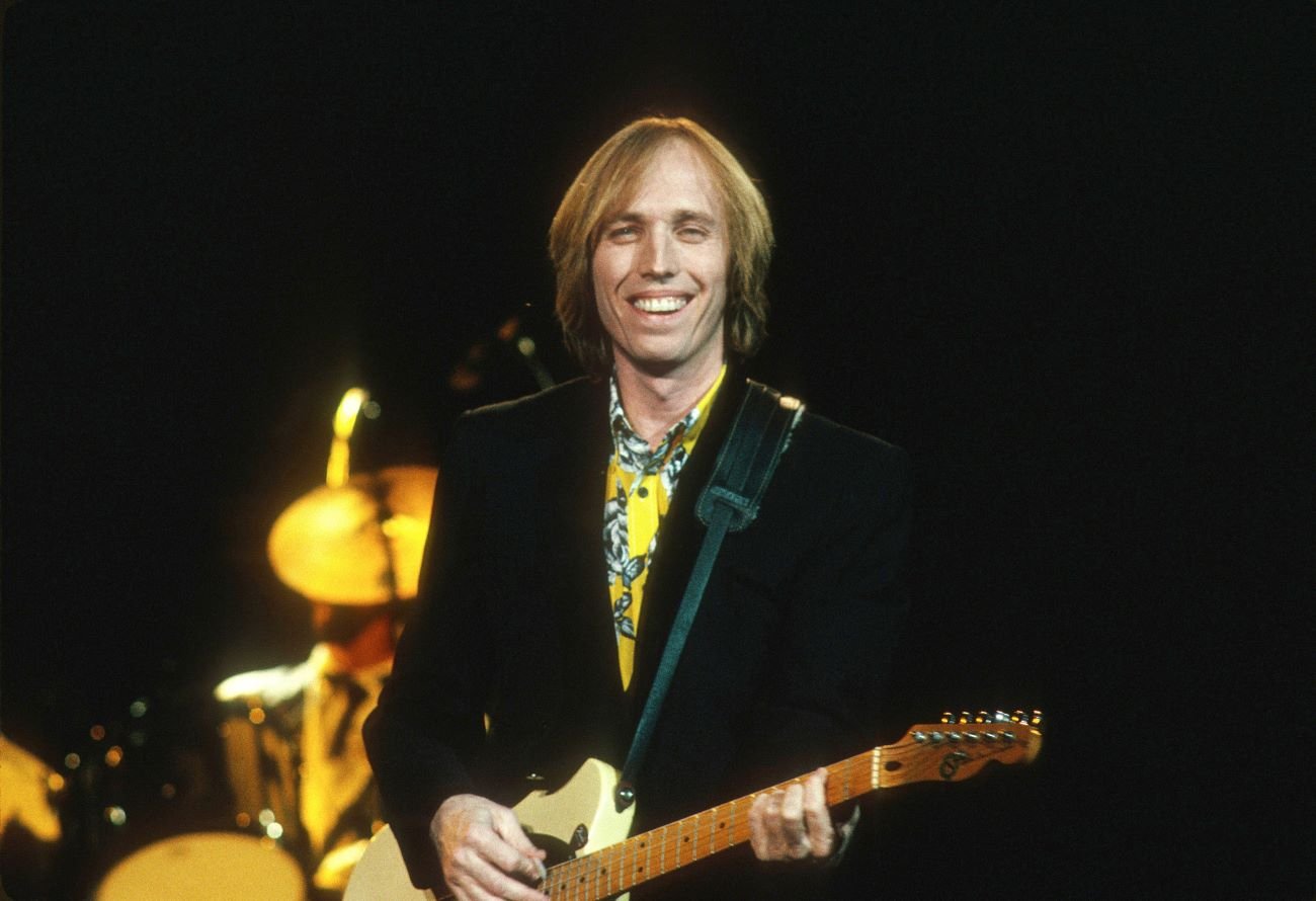 Tom Petty wears a yellow shirt and jacket and holds a guitar.