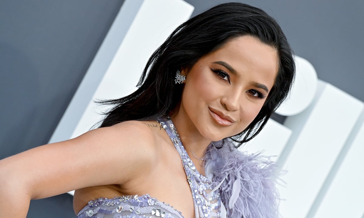 Becky G wears a purple gown with feathers and poses with a smile. 