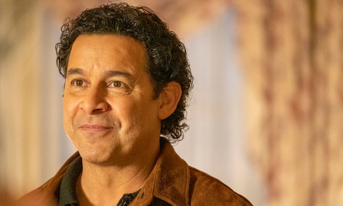 Jon Huertas as Miguel in This is Us wears a brown jacket and smiles.