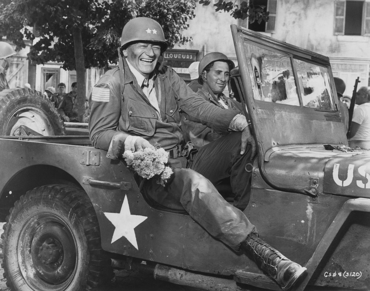 Actor John Wayne, who refused to serve in the military, on the set of 'Cast a Giant Shadow' with his leg hanging out the side of a military vehicle.