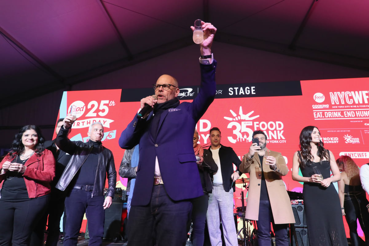 Alton Brown wears a blue blazer and scarf as he raises a glass at a Food Network event.