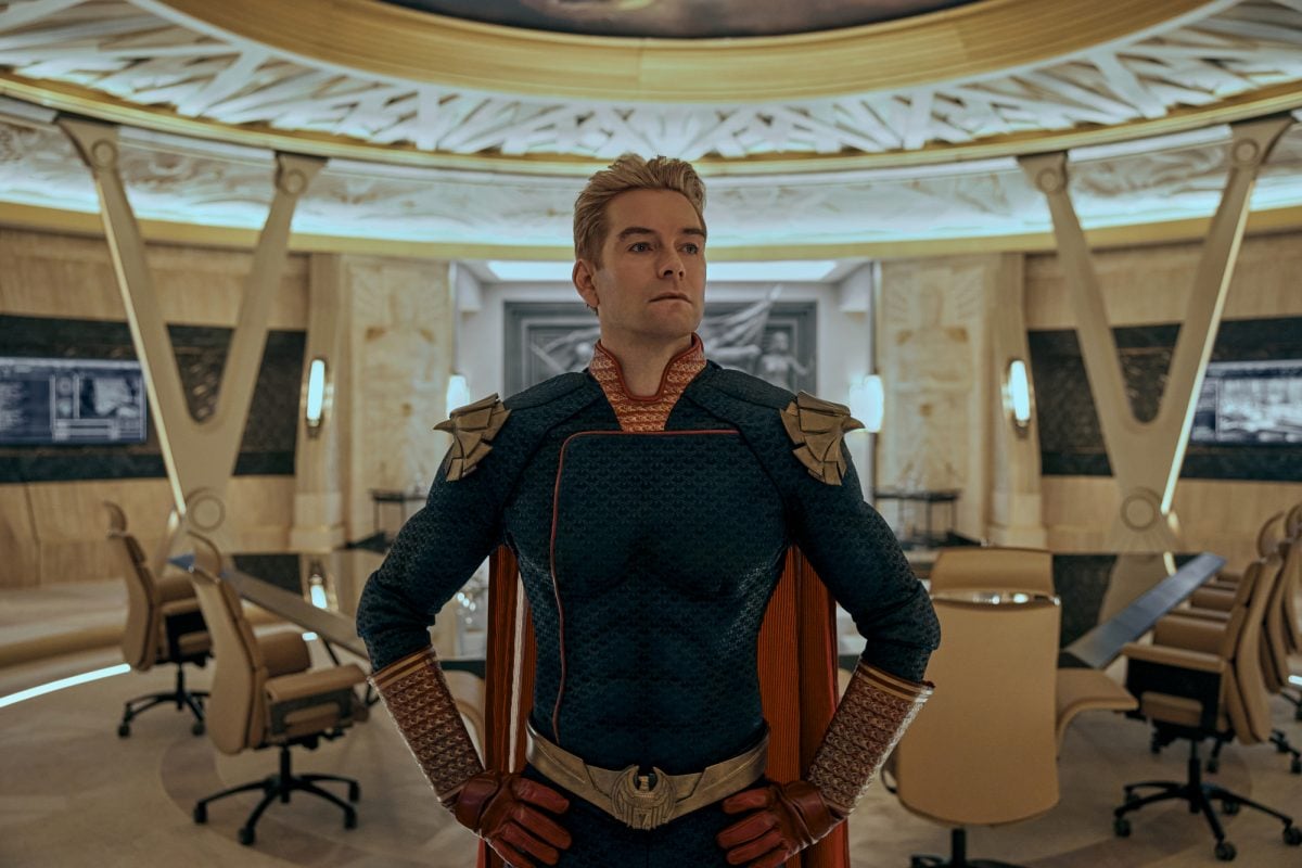 Antony Starr as Homelander in 'The Boys' Season 3. He's wearing his cape and standing in front of a conference room.