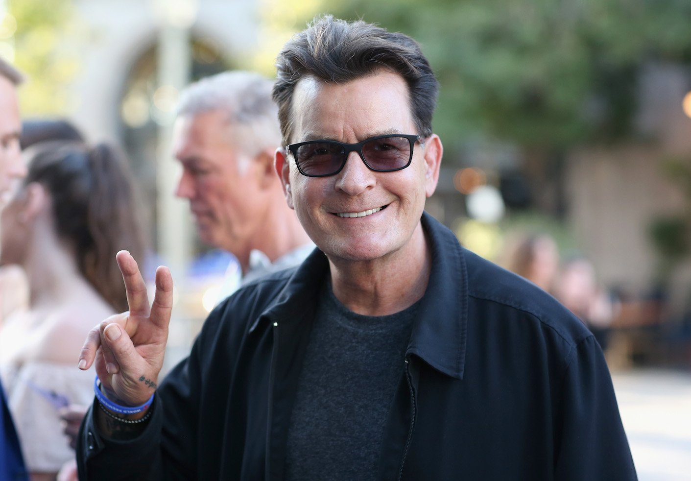 Charlie Sheen smiling while wearing sunglasses and holding up a peace sign with his fingers