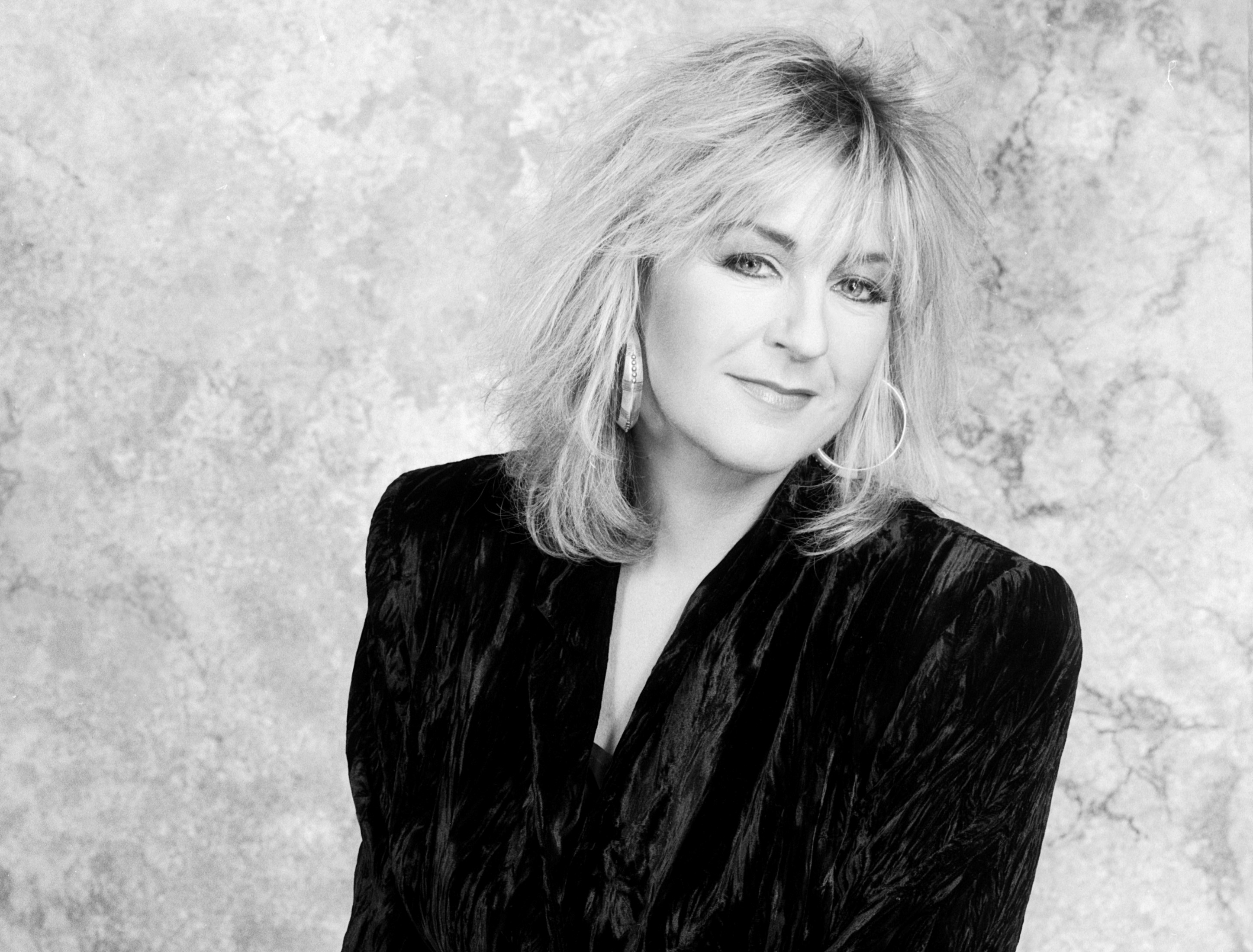 A black and white picture of Christine McVie wearing a blazer and hoop earrings.