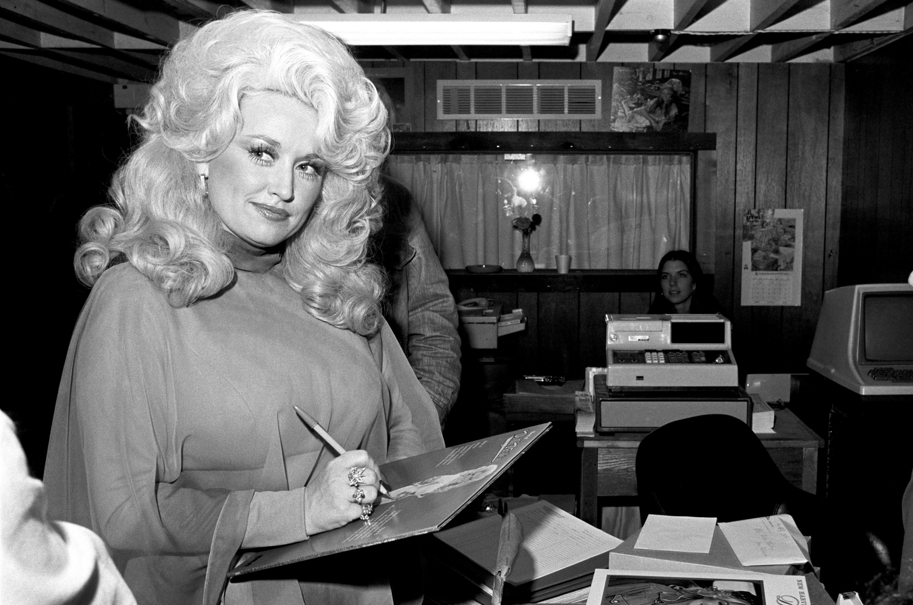 Dolly Parton is in the middle of writing her name on an album. She wears a turtleneck.