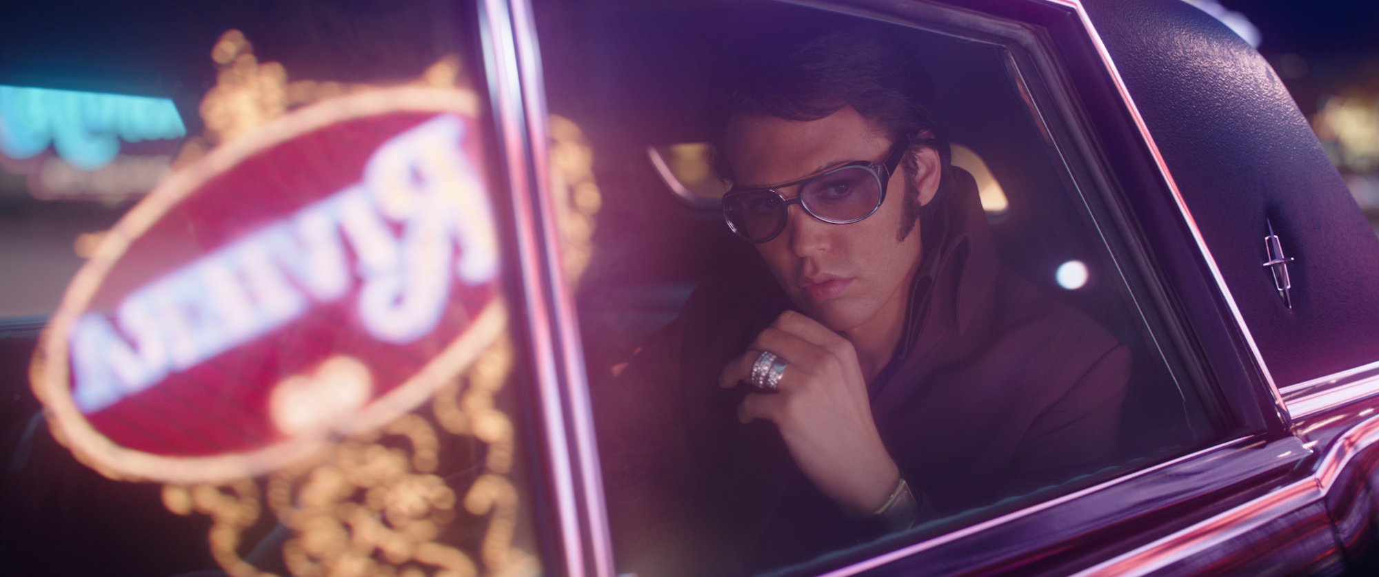 'Elvis' Austin Butler as Elvis Presley wearing sunglasses looking out of the car window with a neon sign reflected in the glass