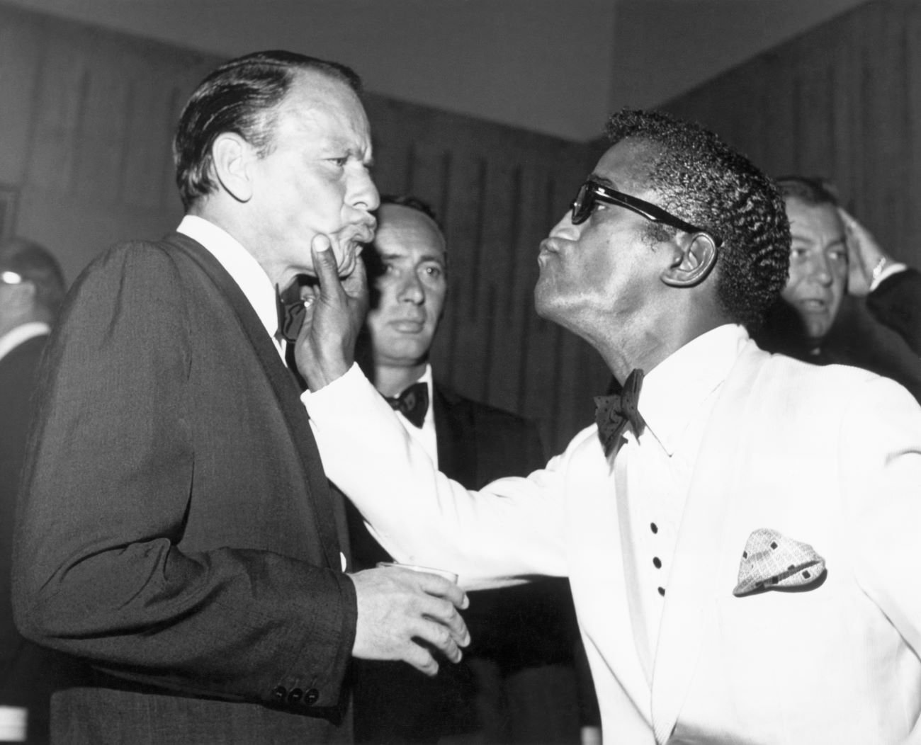 A black and white picture of Sammy Davis Jr. grabbing Frank Sinatra's cheeks. Both men have their lips pursed.