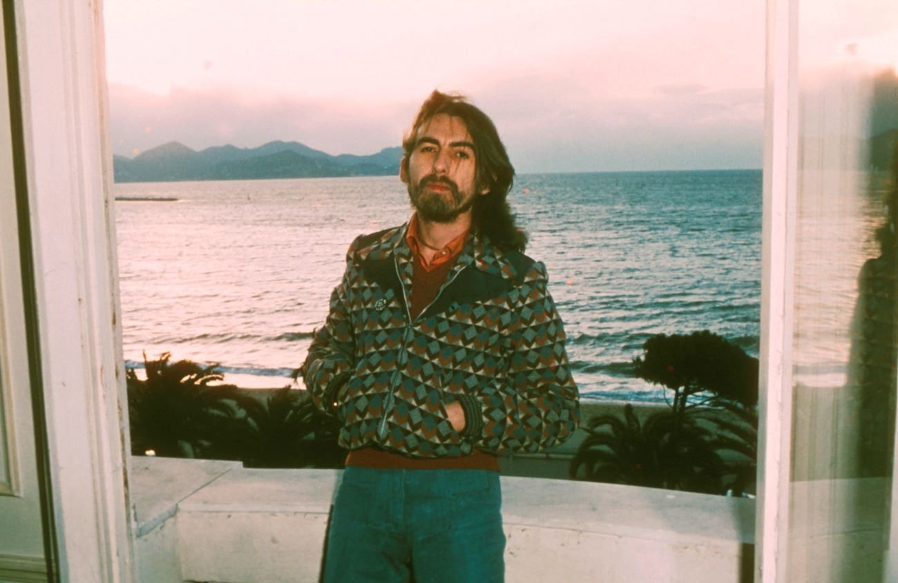 George Harrison stands with his hands in his pockets in front of the ocean.