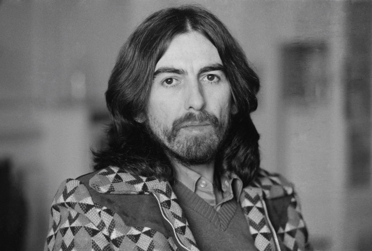 A black and white picture of George Harrison wearing a sweater and a jacket.