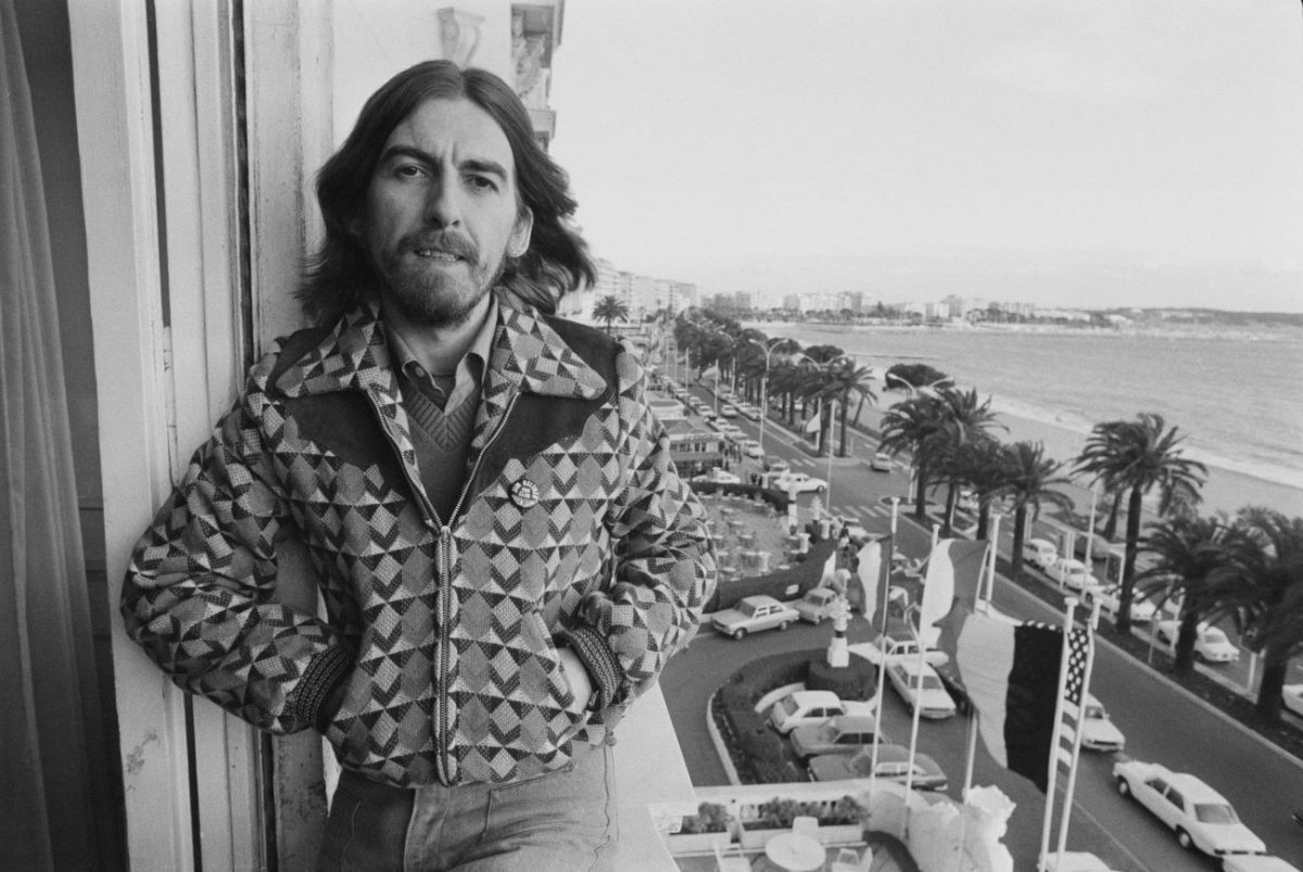 George Harrison stands on a balcony with his hands in his jacket pockets.